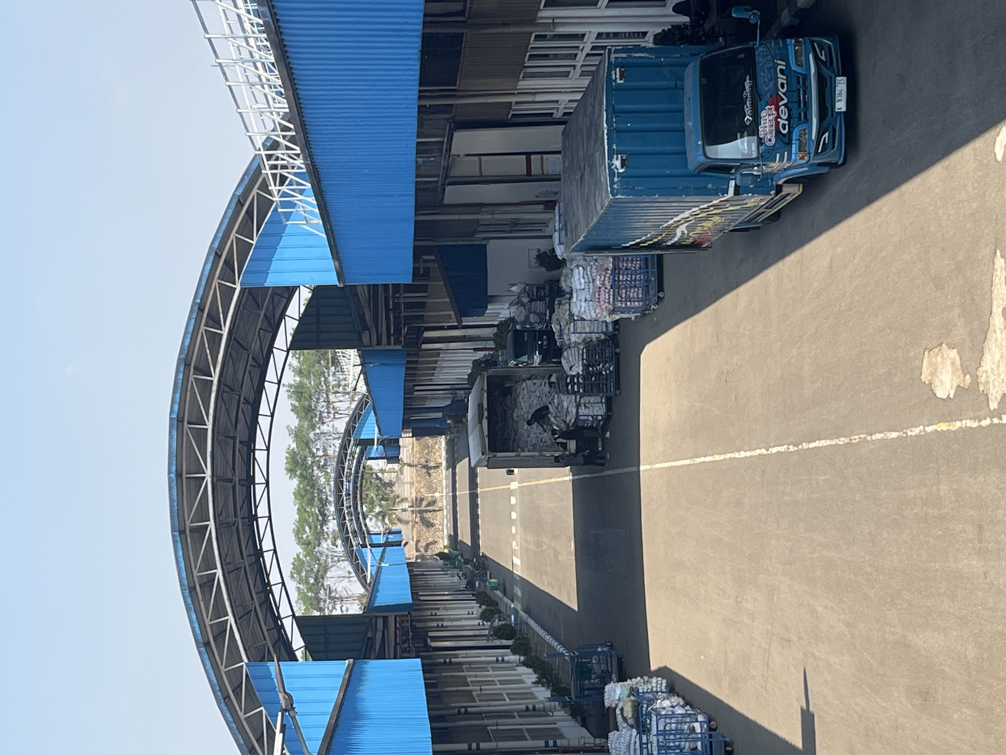 An aerial view captures a logistics facility under a bright, clear sky, suggesting a sunny day in Pelang, Indonesia. The scene depicts a long, asphalt-paved loading area between two parallel rows of industrial buildings. These buildings feature bright blue corrugated metal siding and extended, arched, blue-roofed overhangs supported by a metal framework.
Along the sides of the loading area, numerous blue metal cages or carts are visible, many filled with white sacks or packages. Several people are engaged in loading or unloading activities. In the middle ground, a white truck is parked, with individuals working around its open rear, moving sacks from the truck onto trolleys. Further down the lane, a blue truck with a container body is parked on the right side. The truck displays the branding "Blue Concept devani" and a license plate reading "K 8384 EC".
The loading area is demarcated by white and yellow lines painted on the asphalt. Trees are visible in the distance, beyond the facility's structures, with some appearing to be palm trees. The overall impression is one of an active, open-air logistics or distribution center.