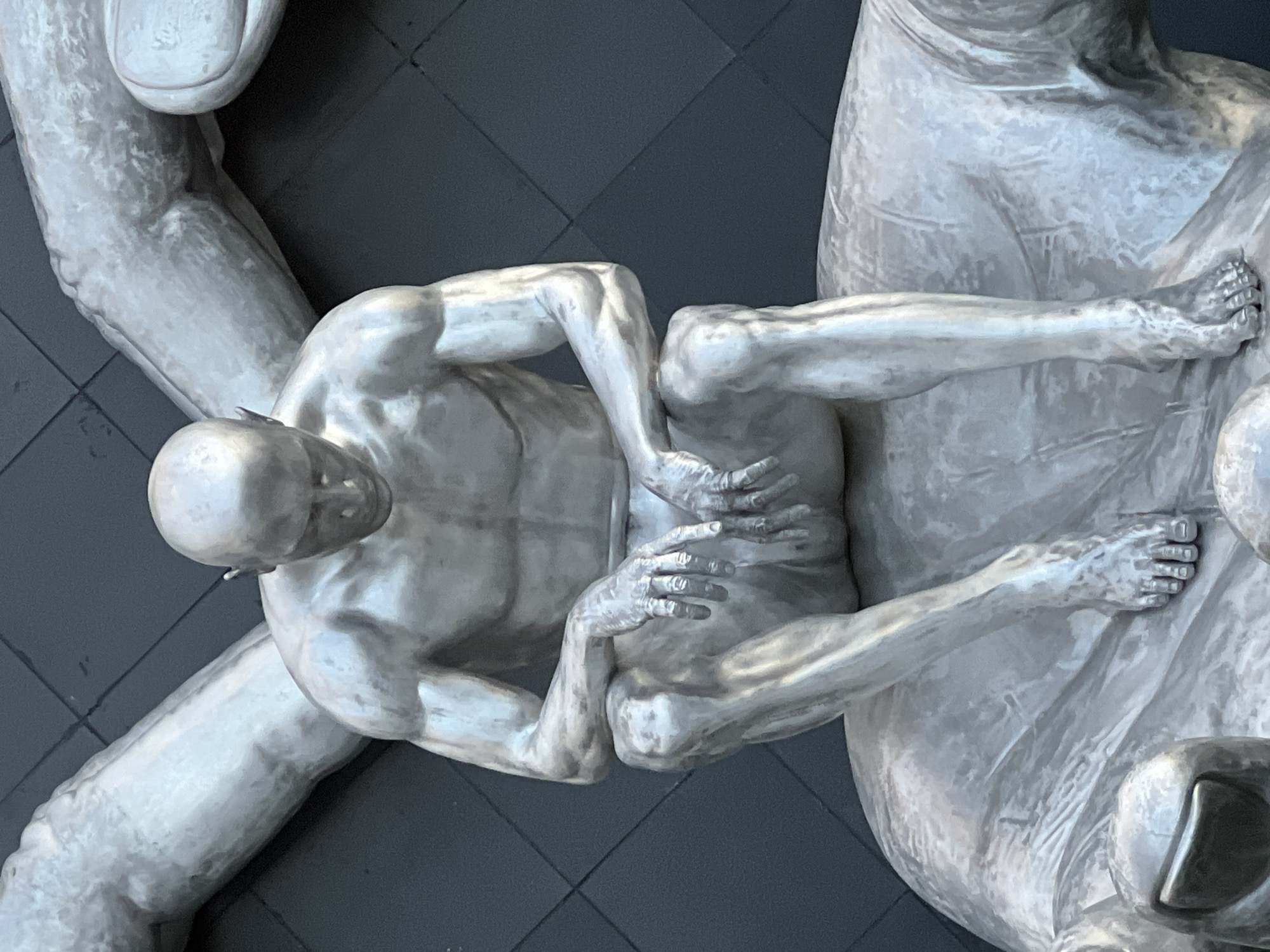 The image shows a large, silver-colored sculpture of a bald man sitting with his head bowed and hands resting on his knees. He appears to be made of metal and has a muscular physique. The sculpture is seated on what appears to be a larger, also silver-colored, sculpture of hands. In the background, a dark, textured wall with a grid pattern is visible. The sculpture is located in Krasnodar, Russia, specifically on V.I. Lenin Street, near the "Avrora" cinema. It is part of the "The Man Who Can't Be Moved" art installation, created by sculptor Sergey Kolpachenko.