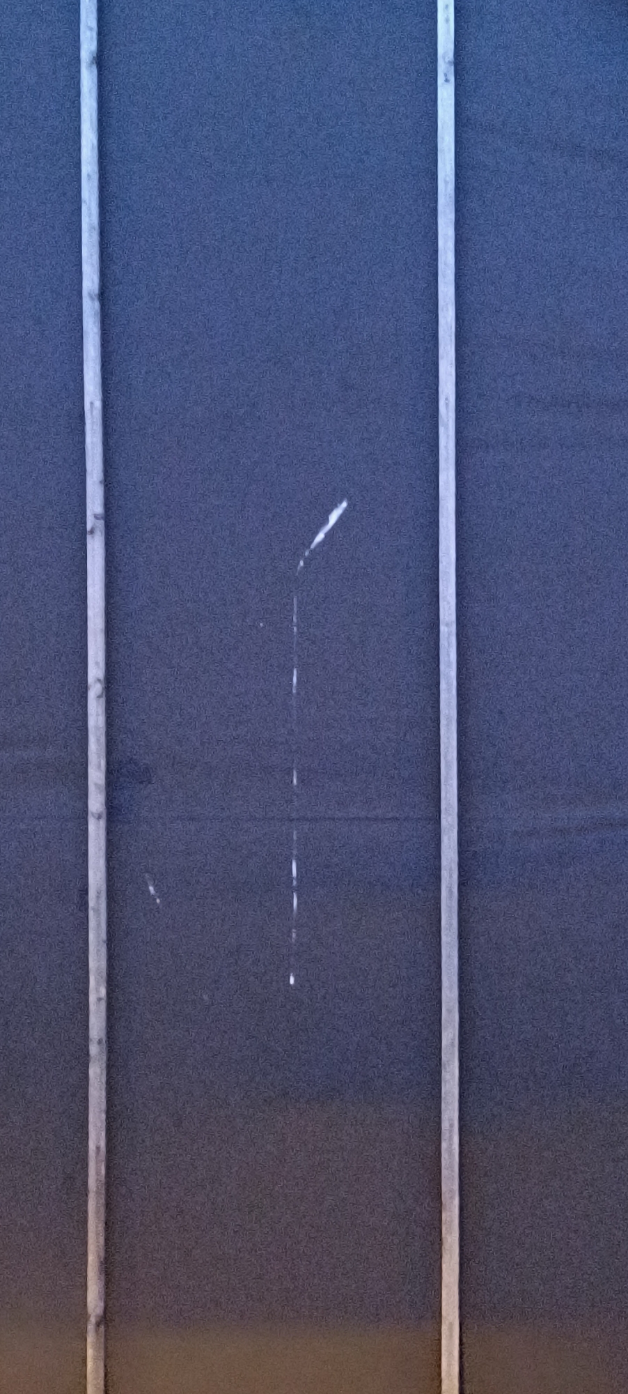 This is a vertical, close-up photograph of a dark blue, textured wall. Two vertical, light-colored, textured wooden beams run parallel to each other, framing the center of the image. A thin, vertical white streak is visible in the center of the frame, appearing to be a drip or stain. This streak has a slight curve at its upper end. A small, faint white mark is also visible to the left of the central streak. The image is grainy, suggesting it was taken in low light conditions. The scene appears to be an exterior wall of a building or structure, likely in an urban or industrial setting, consistent with being located in the city of Winnenden, Germany. There are no people or other discernible subjects in the frame.