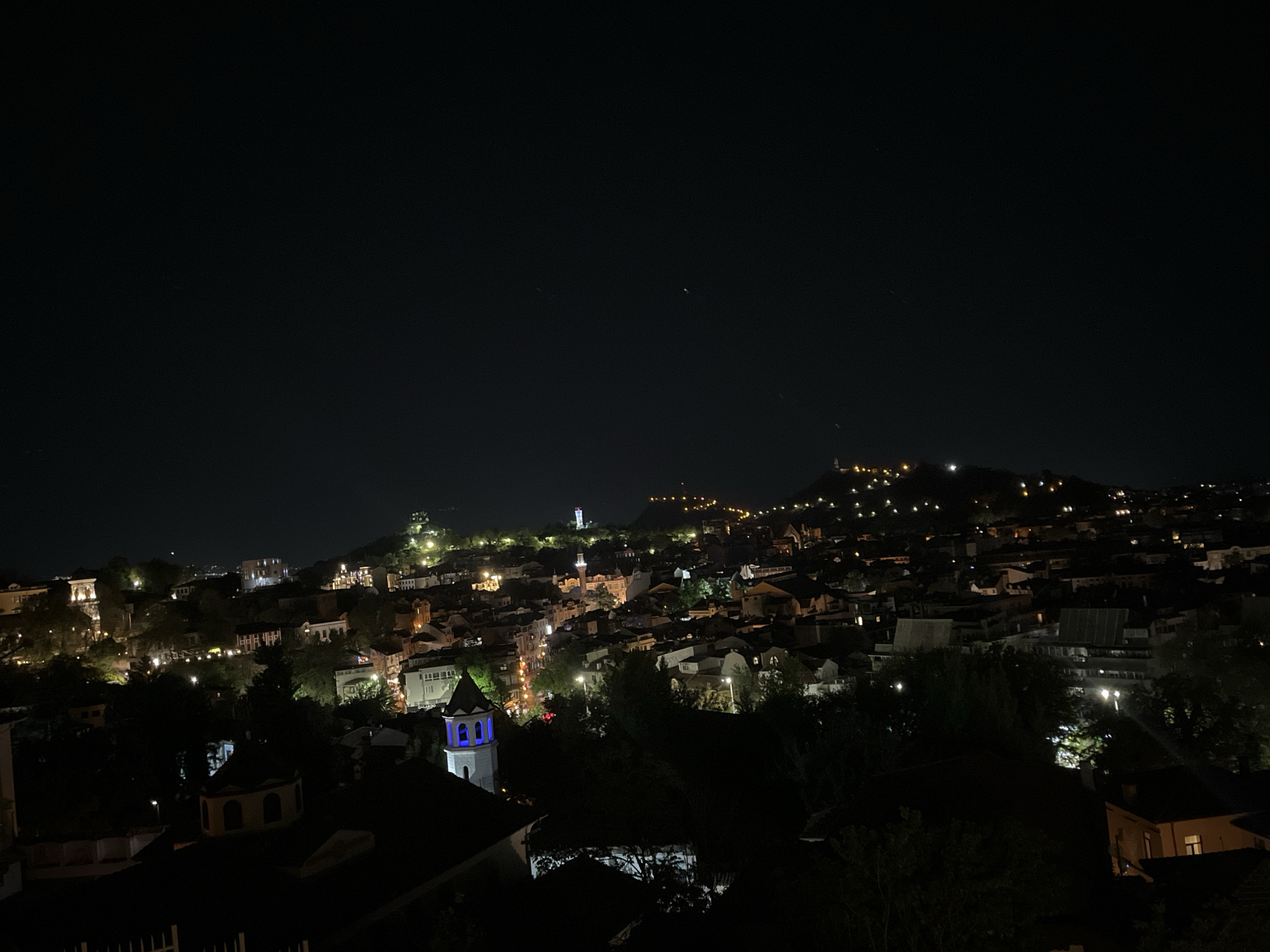At night, a high-angle view shows the city of Plovdiv, Bulgaria. A hilltop is illuminated with numerous small lights. A building with a tower with a blue illuminated facade is in the foreground. The sky is dark.