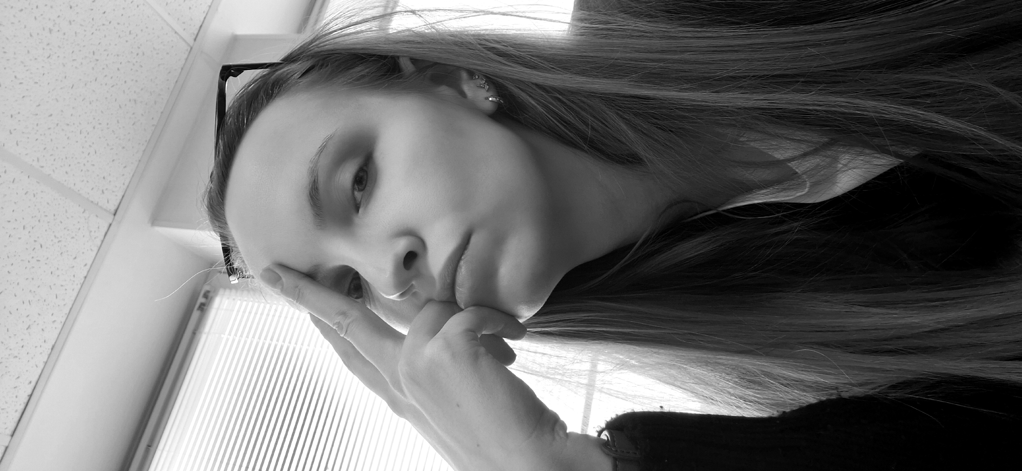 A black and white, tilted, medium shot depicts a young woman indoors. She rests her forehead on her right index finger, with the rest of her hand obscuring part of her face. She has long hair, parted to one side, and glasses are perched on her forehead. Her head is angled downwards. The background includes a window with blinds and a ceiling with a grid pattern. The location of the image cannot be determined beyond it being an indoor location. The city of Chaikovsky, Russia cannot be verified from the image.