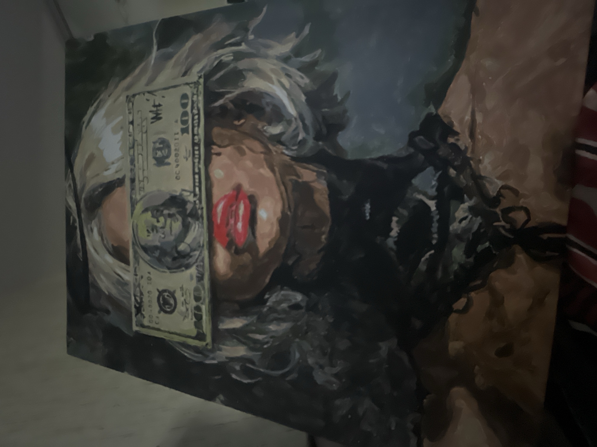 The image displays a photograph of a painting featuring a stylized portrait of a person, likely a woman, with light-colored, textured hair. Her eyes are entirely obscured by a vertical hundred-dollar bill placed across her face. Below the bill, her mouth is visible, characterized by vibrantly red lips. The background of the painting is dark, appearing in shades of deep blue and gray. To the right of the painted figure, indistinct dark shapes suggest part of her body or clothing.
The hundred-dollar bill itself is detailed, showing the number "100" in several places and a portrait resembling Benjamin Franklin. Closer examination of the bill reveals non-standard text markings such as "WF-", "GC 9002011", and "00-8020 10A", suggesting it may be a prop or an artistic interpretation rather than an authentic US banknote. The painting's composition, with the eyes covered by money, conveys potential themes related to wealth, blindness, censorship, or perception. The photograph is taken indoors, with a plain, light-colored wall visible to the left and a blurred reddish-brown object to the right, offering minimal context about the physical setting.