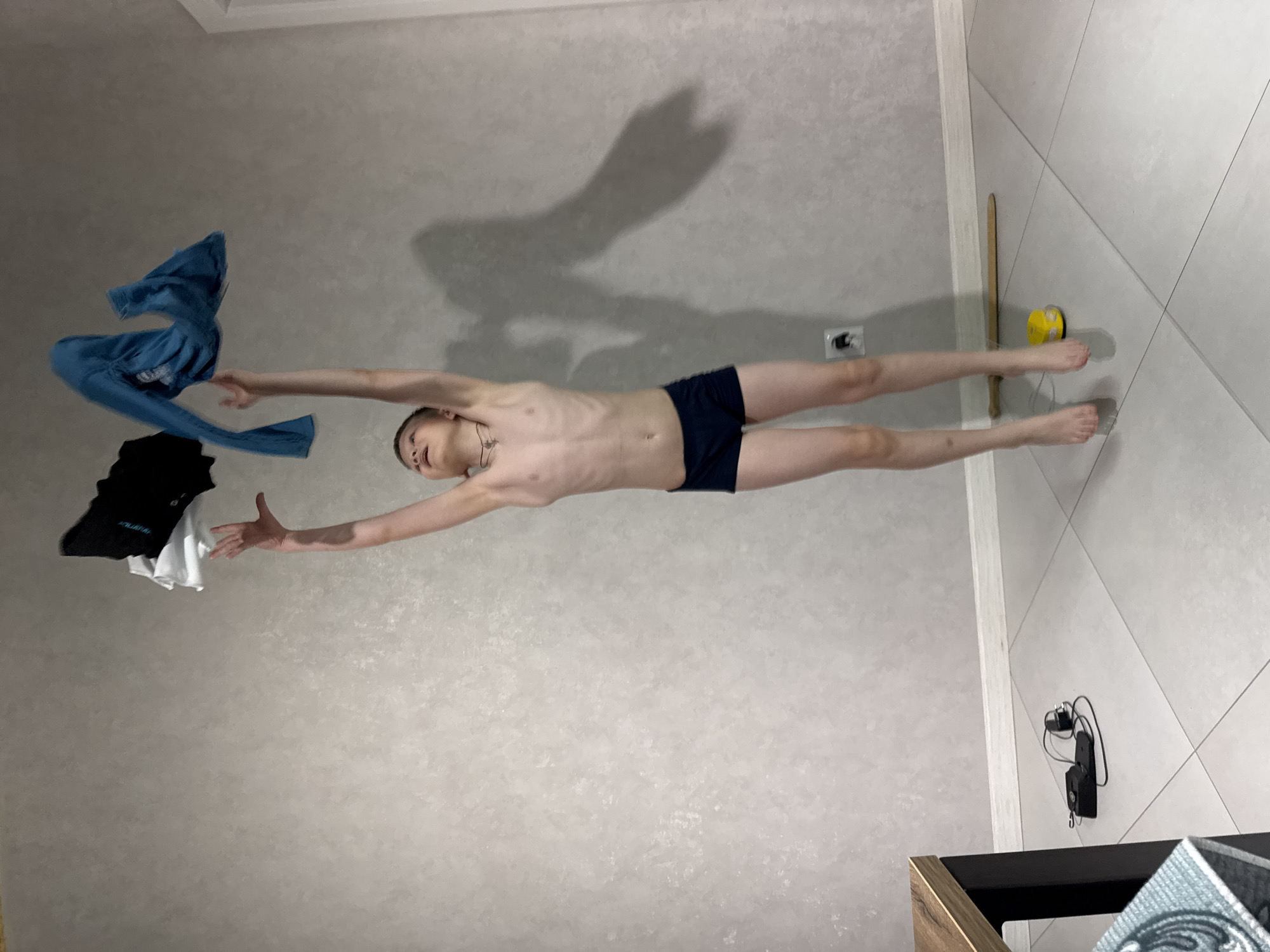 A shirtless young man in dark underwear is lying horizontally on the floor of a room in Gornyi (Dvurechenskoe S/P), Russia, with his arms outstretched, appearing to simulate flight. Items of clothing (blue shirt, black shirt, and white shirt) are suspended near his left hand, as if being caught or dropped. His shadow is cast on the wall above him. To the right, a small yellow object lies on the floor near a stick-like object. Further down the wall, a black electrical device is mounted on the wall near the floor. The floor is tiled.