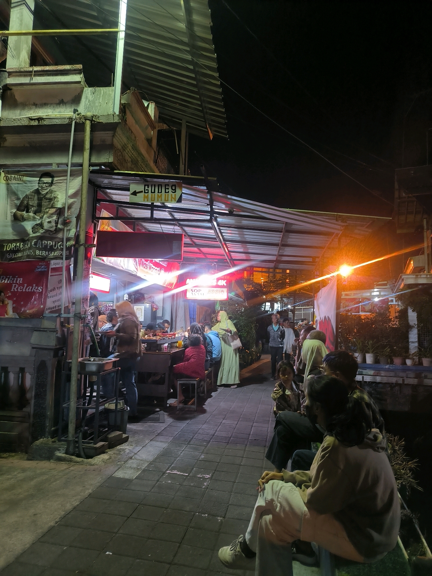 The image captures a nighttime street scene in Salatiga, Indonesia, specifically in front of a food stall area. The foreground shows a tiled walkway with several people seated on low benches, facing a brightly lit food stall. In the middle ground, patrons are seated at tables within the covered food stall, eating and socializing. A woman in a hijab is walking away from the camera towards the stall. Behind the food stalls, there is a dark alleyway with some potted plants. The overall atmosphere is lively and communal, typical of a street food scene at night. The architecture suggests a simple, functional design for the food stalls, with corrugated metal roofing and awnings. There are advertisements and signs displayed on the buildings and stalls. The lighting is a mix of warm artificial lights from the stalls and ambient streetlights, creating a somewhat hazy and atmospheric effect. The sky is dark, indicating it is well past sunset.