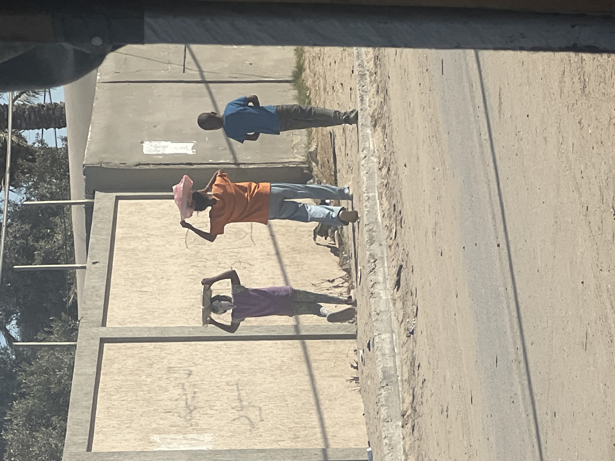 In Deir el-Balah, Palestinian Territories, three individuals are walking along a dirt path next to a paved road. The person on the left, wearing a purple shirt and grey pants, carries a flat, round object on their head. The middle person, in an orange t-shirt and blue pants, also has a pink, bowl-shaped object balanced on their head. The person on the right, dressed in a blue t-shirt and grey pants, walks without carrying anything. Behind them is a textured, light-colored wall, and beyond that, trees and a clear sky are visible. The ground is dusty with scattered debris.