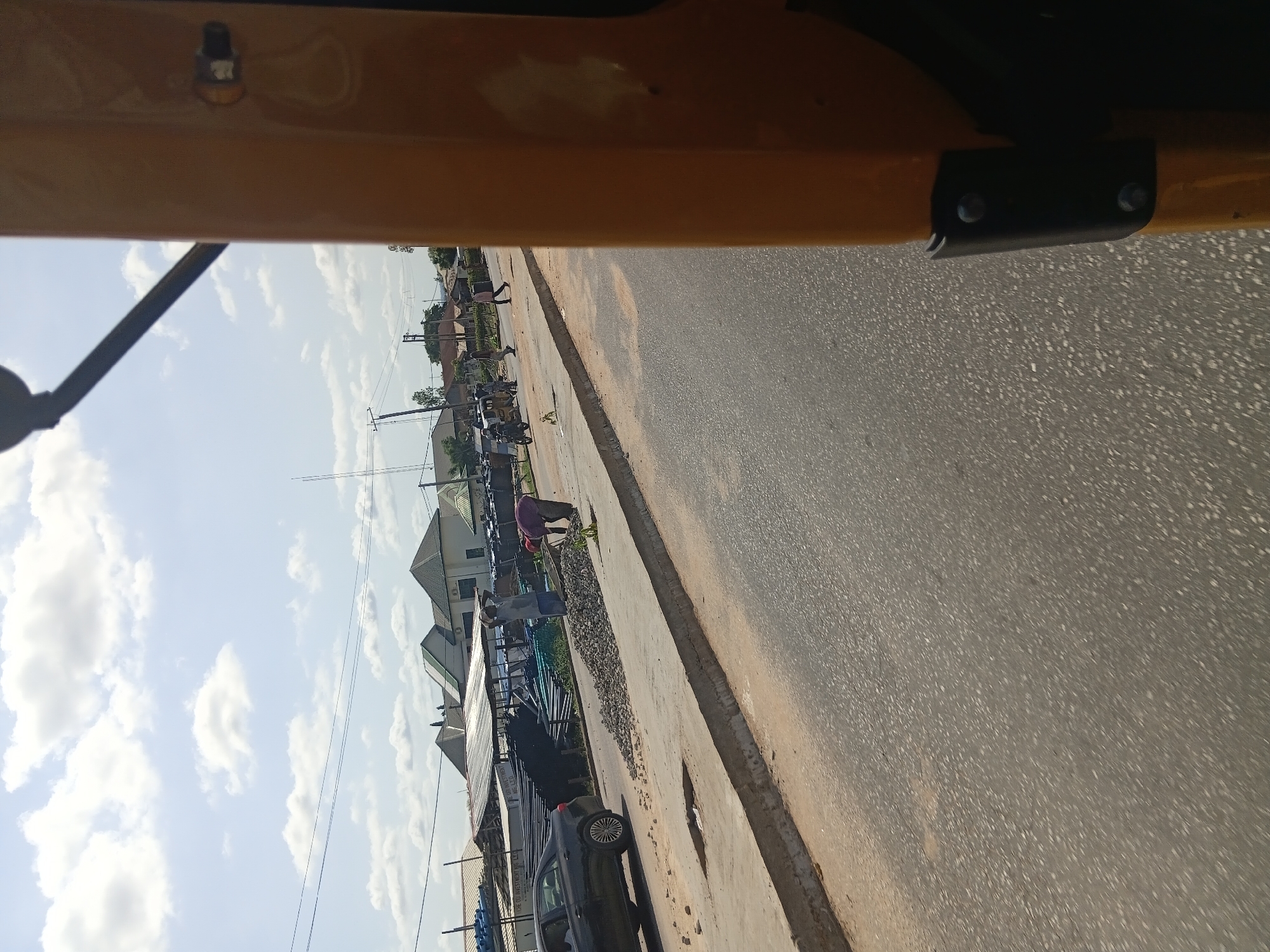 The image depicts a street scene in Jalingo, Nigeria. The perspective suggests the photograph was taken from inside a vehicle, possibly a tuk-tuk, given the yellow overhead structure visible in the foreground. The scene is dominated by a paved road on the right and a sidewalk or cleared area on the left, lined with buildings and some scattered debris.
Several people are visible. On the sidewalk, one person in a purple shirt is bending down, possibly engaged in some task. Further down the sidewalk, another person, dressed in blue and gray, is also stooping. A line of parked motorcycles or bicycles is also present along the sidewalk. Behind these, a black car is parked on the side of the road.
The buildings in the background appear to be residential or commercial, with tiled roofs and various architectural styles. Power lines are visible stretching across the scene. The sky is bright with scattered white clouds, indicating it is daytime, likely morning or afternoon, with good weather.
Notable details include the rough texture of the road and sidewalk, the presence of construction materials like gravel and pipes, and the overall bustling atmosphere suggested by the activity and parked vehicles. The image provides a glimpse into daily life on a street in this Nigerian city. No visible text is decipherable.