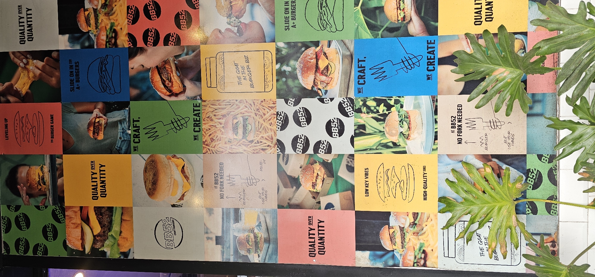 The image shows a wall covered in square panels, each featuring different images and text related to a burger restaurant named "BB52," presumably located in the Ubud District of Indonesia. The panels display a variety of designs including photographs of burgers, graphic illustrations related to burgers, the restaurant's branding, and text slogans like "Quality Over Quantity," "Slide on in for A+ Burgers," and "No Fork Needed." Some panels show parts of people eating burgers. Some of the panels are obscured by green leafy plant.