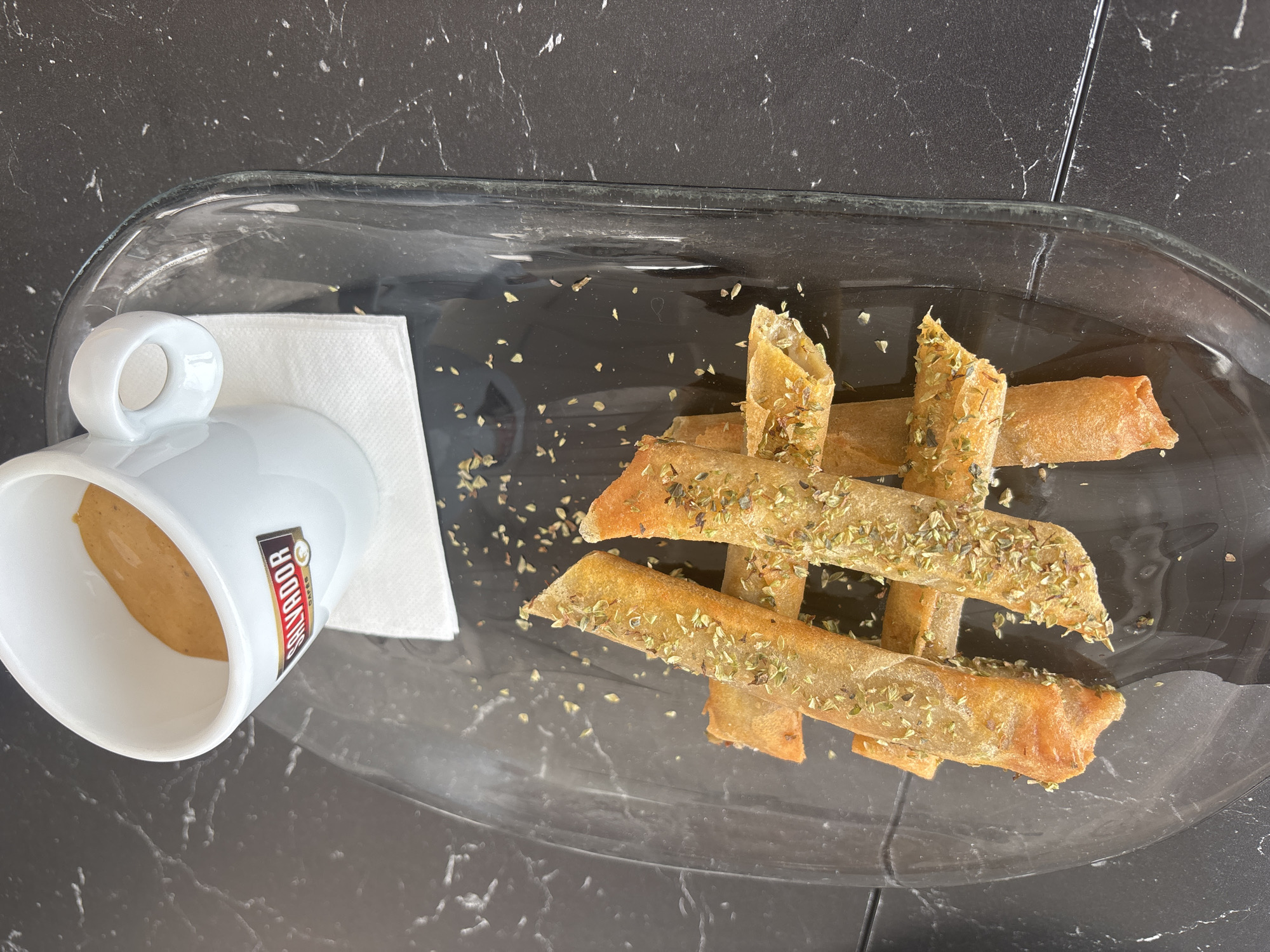 A white espresso cup, bearing the "SALVADOR CAFES" logo, sits on a white paper napkin. The cup is filled with a light brown liquid, likely coffee or a similar beverage. Beside the cup, on a clear glass rectangular dish, are several golden-brown pastry rolls sprinkled with dried herbs. The dish rests on a dark, marbled surface, possibly a table. The scene appears to be an indoor setting, likely a cafe or restaurant in El Parador de las Hortichuelas, Spain. No people are visible in the image. The lighting is overhead, casting subtle shadows. The overall presentation is simple and focused on the food and drink.