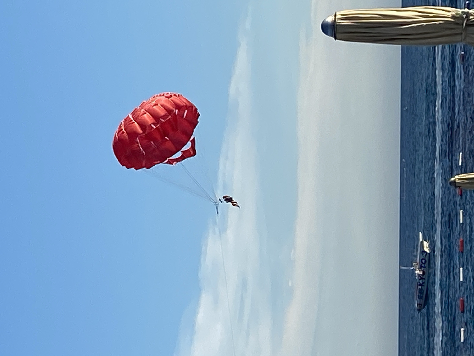A red parachute with two people parasailing is visible against a bright blue sky. Below, the blue sea is calm with whitecaps. In the distance, a white boat with "KYOTO 3" written on its side is sailing. In the foreground, a closed beach umbrella stands on the right side of the image, and buoys are visible in the water. The scene appears to be a coastal area in Budva, Montenegro.