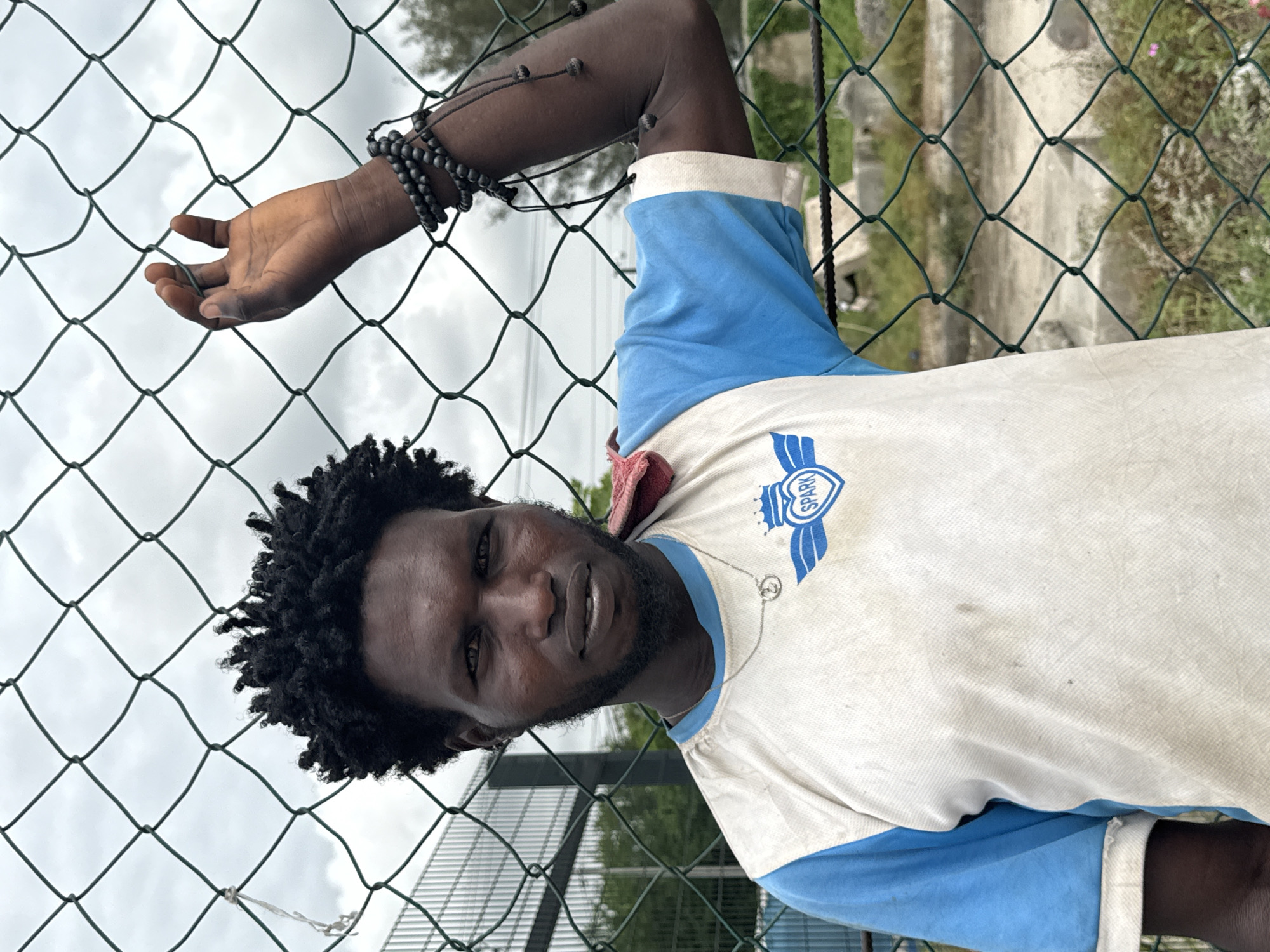 A Black man with a beard and dreadlocks is shown from the chest up, leaning against a green wire fence. He is wearing a t-shirt with blue sleeves and a white front that has a logo on it that reads "SPARK." The man is wearing bracelets on his left wrist and on his left upper arm. He has his left hand extended towards the fence. The background shows greenery and an overcast sky. This photo was taken near the city of Lekki, Nigeria.