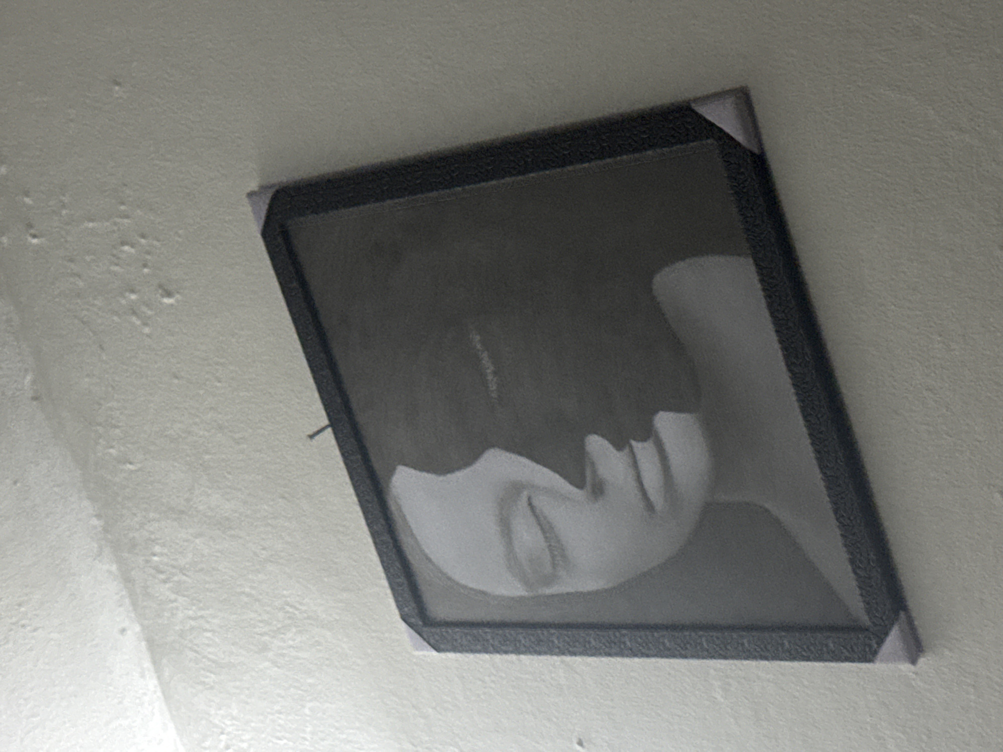 A framed black-and-white picture hangs askew on a light-colored wall. The picture depicts a person with dark hair lying down. The location is unknown, but the request included Ihiagwa, Nigeria. There is peeling paint visible on the wall to the left of the picture. The frame has gray corner protectors. A thin nail is visible next to the frame.