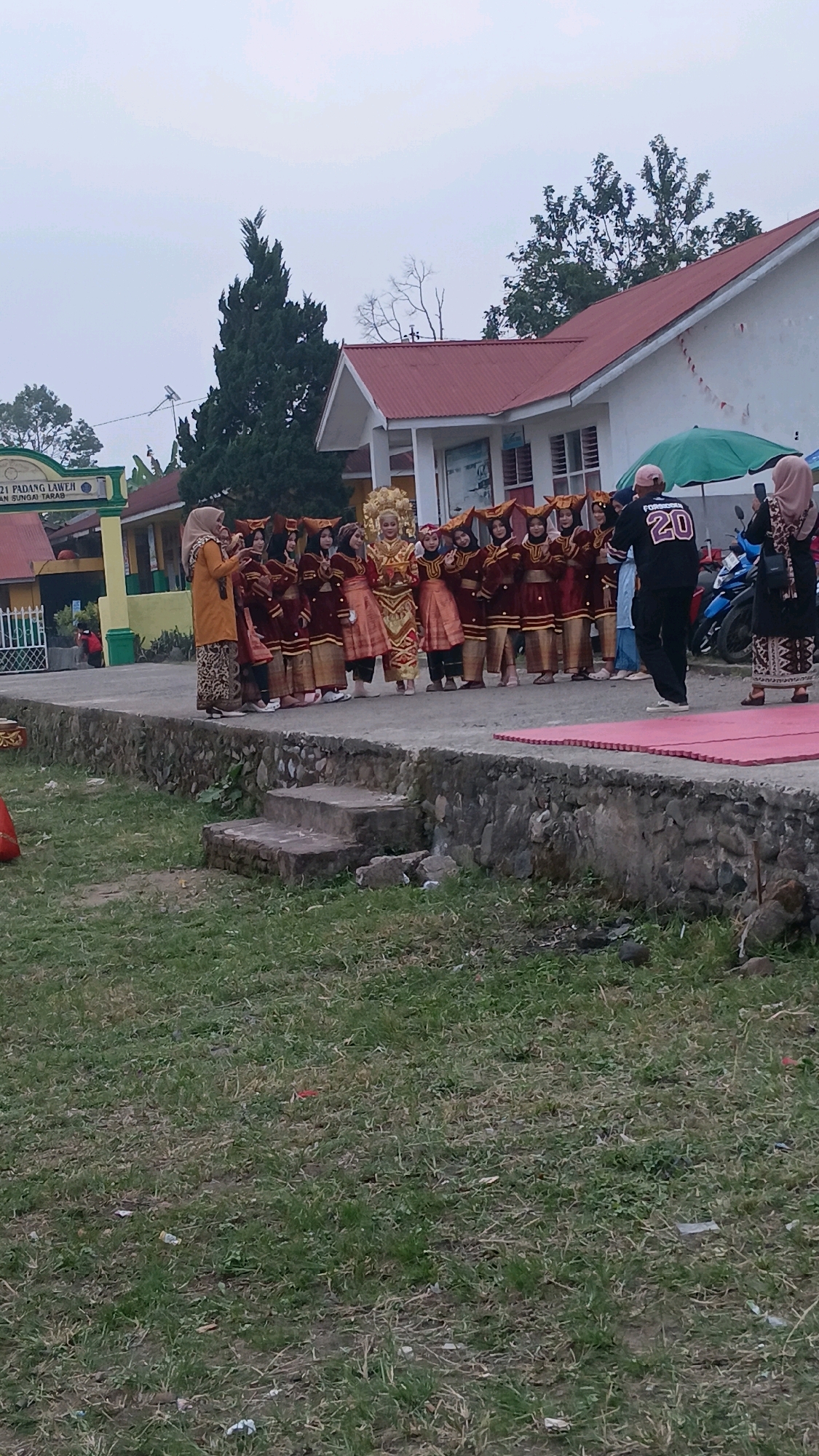 In Padang Laweh, Indonesia, a group of individuals, primarily young women in traditional attire, are gathered outside a building with a red-tiled roof. The women are dressed in matching maroon and gold outfits, some with elaborate headwear. One central figure wears a more ornate, golden costume and headpiece, suggesting a performance or special occasion. They are standing in a line on a raised platform, with a stone wall and steps leading down to a grassy area in the foreground. A few adults are present; one person in a black shirt with "20" visible on the back is facing the group, and another individual wearing a headscarf is positioned at the far right of the line, possibly taking a photo. A green umbrella is visible behind the group. The environment appears to be an outdoor area adjacent to a school or community building, indicated by signage visible in the background. The sky is overcast, suggesting it might be early morning or late afternoon.