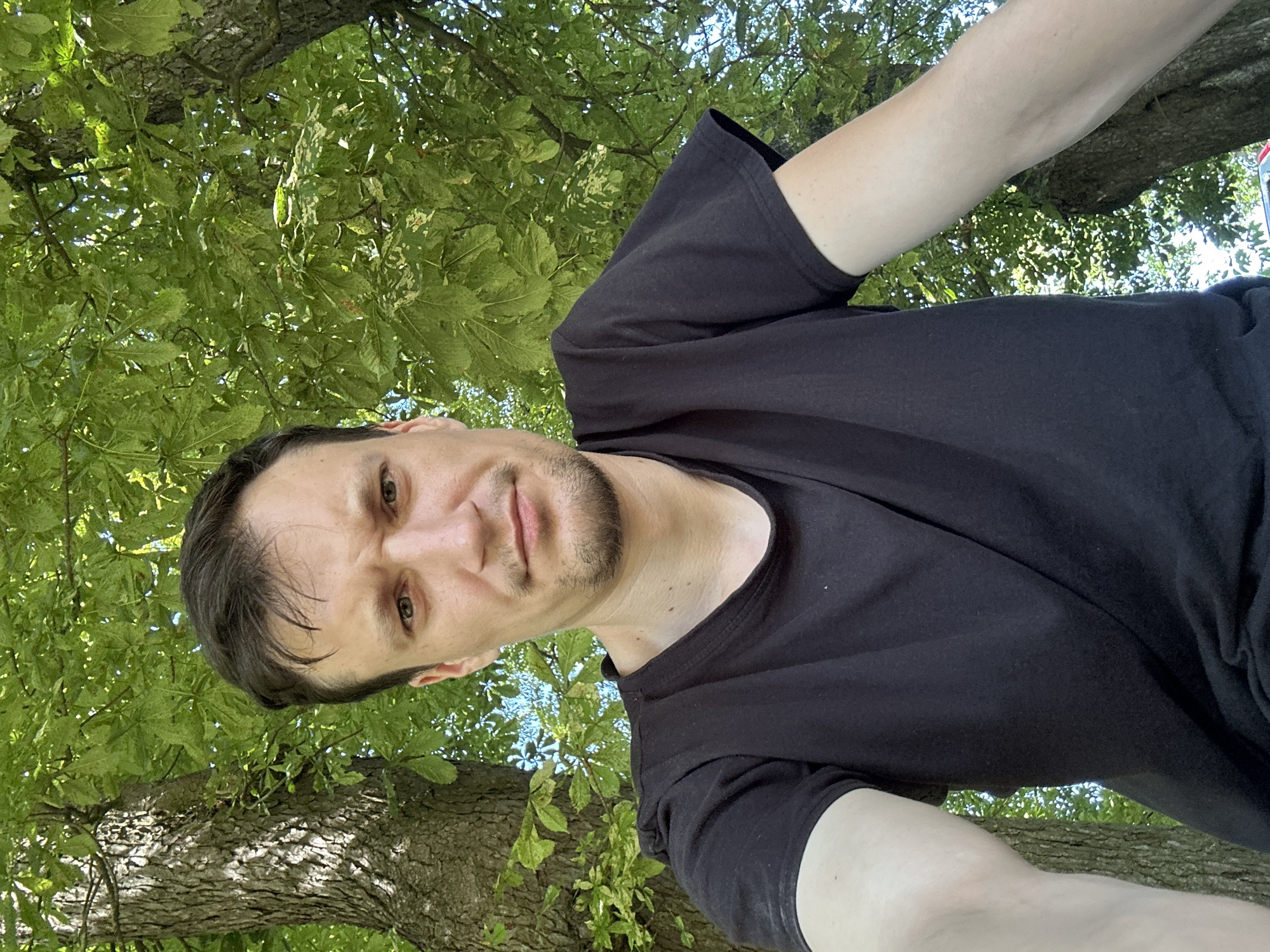 A medium shot shows a man in a black t-shirt standing outdoors under a large tree with lush green foliage. The man has short, dark hair, a light complexion, and a short beard. His facial expression is neutral, with his eyes looking directly at the camera. The background consists of the tree trunk and leaves, with some blue sky visible through the branches. The lighting suggests it is daytime. No specific location in Makiv, Ukraine, can be determined from the image.