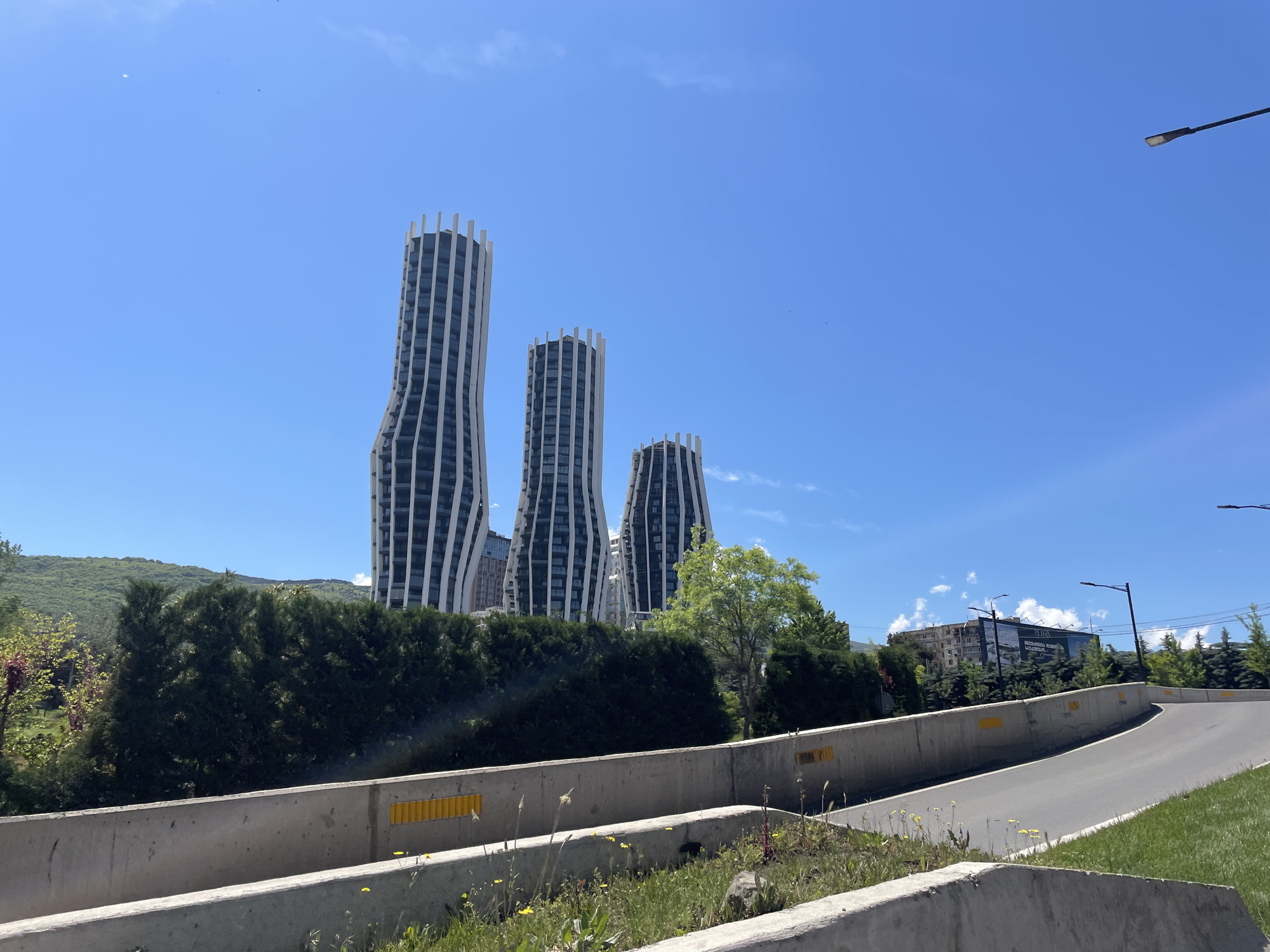 The image shows three modern, high-rise buildings with a distinctive wave-like architectural design, set against a clear blue sky. These buildings are located in the city of Tbilisi, Georgia. In the foreground, there is a concrete retaining wall bordering a road, with some greenery (grass and small flowering plants) growing along the base of the wall. Beyond the retaining wall, there is a row of dark green trees and shrubs, with some buildings and a billboard visible in the background. A street light pole is visible on the right side of the frame. The overall scene suggests a sunny day in a suburban or outskirts area of Tbilisi.