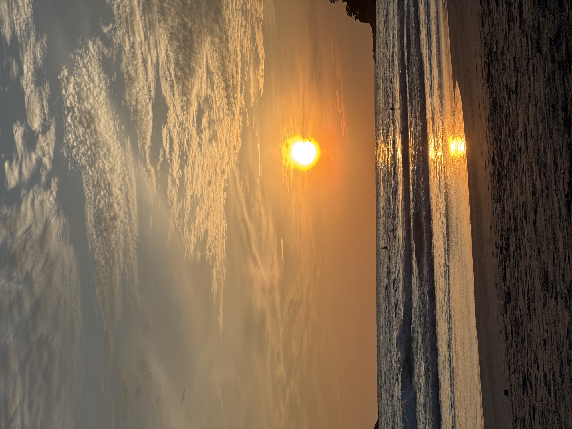 The image depicts a tranquil sunset scene at Karon Beach in Karon, Thailand. The sun, a bright orange orb, is low on the horizon, casting a warm glow across the sky and the ocean. Wispy clouds are scattered across the sky, tinged with orange and yellow hues. Gentle waves roll onto the sandy shore, creating a soft, rhythmic sound. A few individuals are visible in the water, their silhouettes dark against the vibrant backdrop. The beach itself is composed of fine sand, with some debris scattered across its surface. A small, forested headland is visible in the distance on the right side of the frame, adding a touch of natural beauty to the landscape. The overall atmosphere is serene and peaceful, inviting viewers to relax and enjoy the natural spectacle of a tropical sunset.