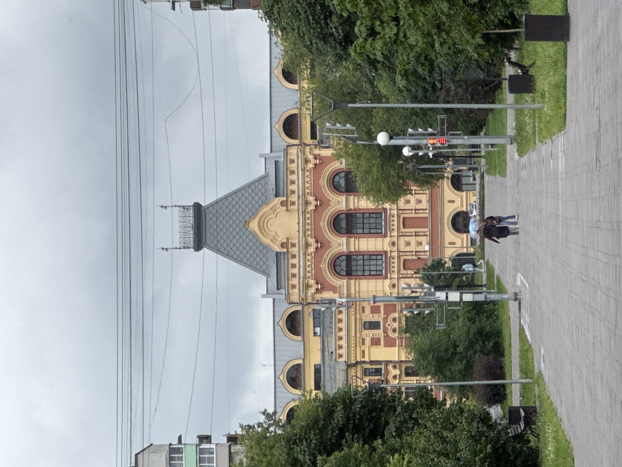 The image shows a view of the Nizhny Novgorod State Museum of Fine Arts, formerly the Alexander Nevsky Cathedral, located in Nizhny Novgorod, Russia. The grand building is designed in a Neo-Russian style with ornate architectural details and a prominent mansard roof. In the foreground, a paved walkway leads towards the museum, flanked by lush green trees and grass. Two individuals, one holding an umbrella, are walking away from the camera on the walkway. Streetlights and traffic signals are visible along the path. The sky is overcast, suggesting a cloudy day. The overall scene depicts a public space with a historical landmark.