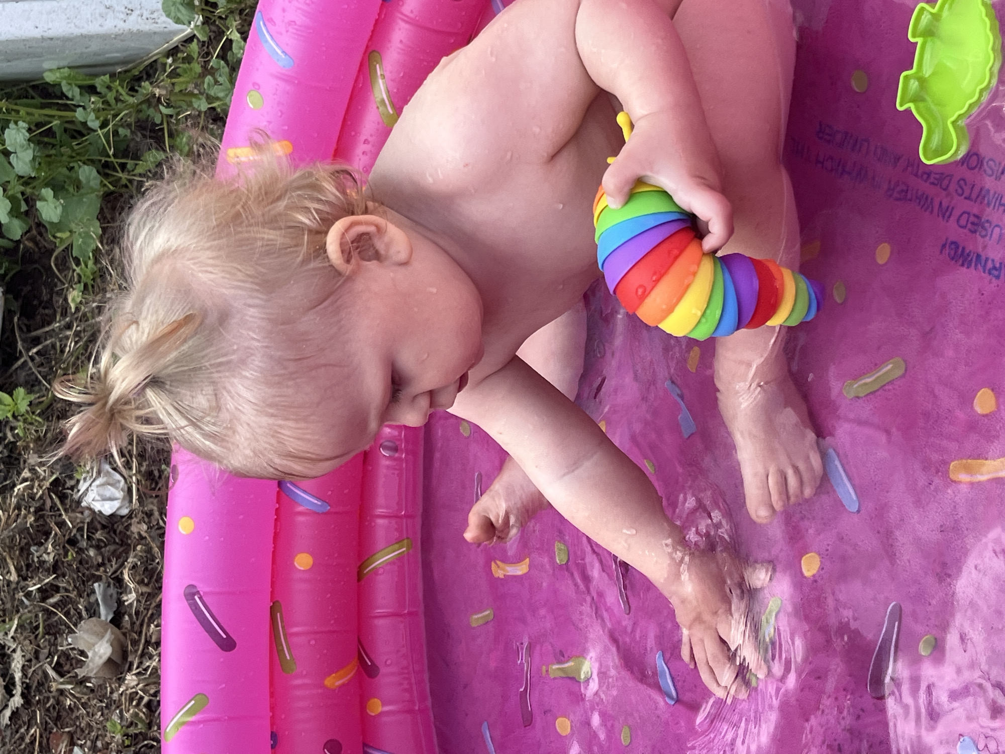 A baby with blonde hair is lying on their side in a pink inflatable pool, possibly a kiddie pool, filled with water. The pool has a sprinkle donut design on its surface. The baby is holding a colorful, segmented caterpillar toy in their right hand. Their skin is wet, indicating they are in the water. The baby's left arm is bent and resting on their body, and their legs are also bent. The setting appears to be outdoors, with dry grass and some greenery visible to the left of the pool, suggesting a backyard or garden. The lighting indicates it is daytime, likely sunny. There is some text printed on the purple surface of the pool, which includes the word "WARNING!" and "USED IN WATER TO WHICH THE", but the rest of the text is not clearly legible. A green plastic toy fish is visible in the upper right corner of the pool. The scene conveys a sense of playful relaxation for the baby.
