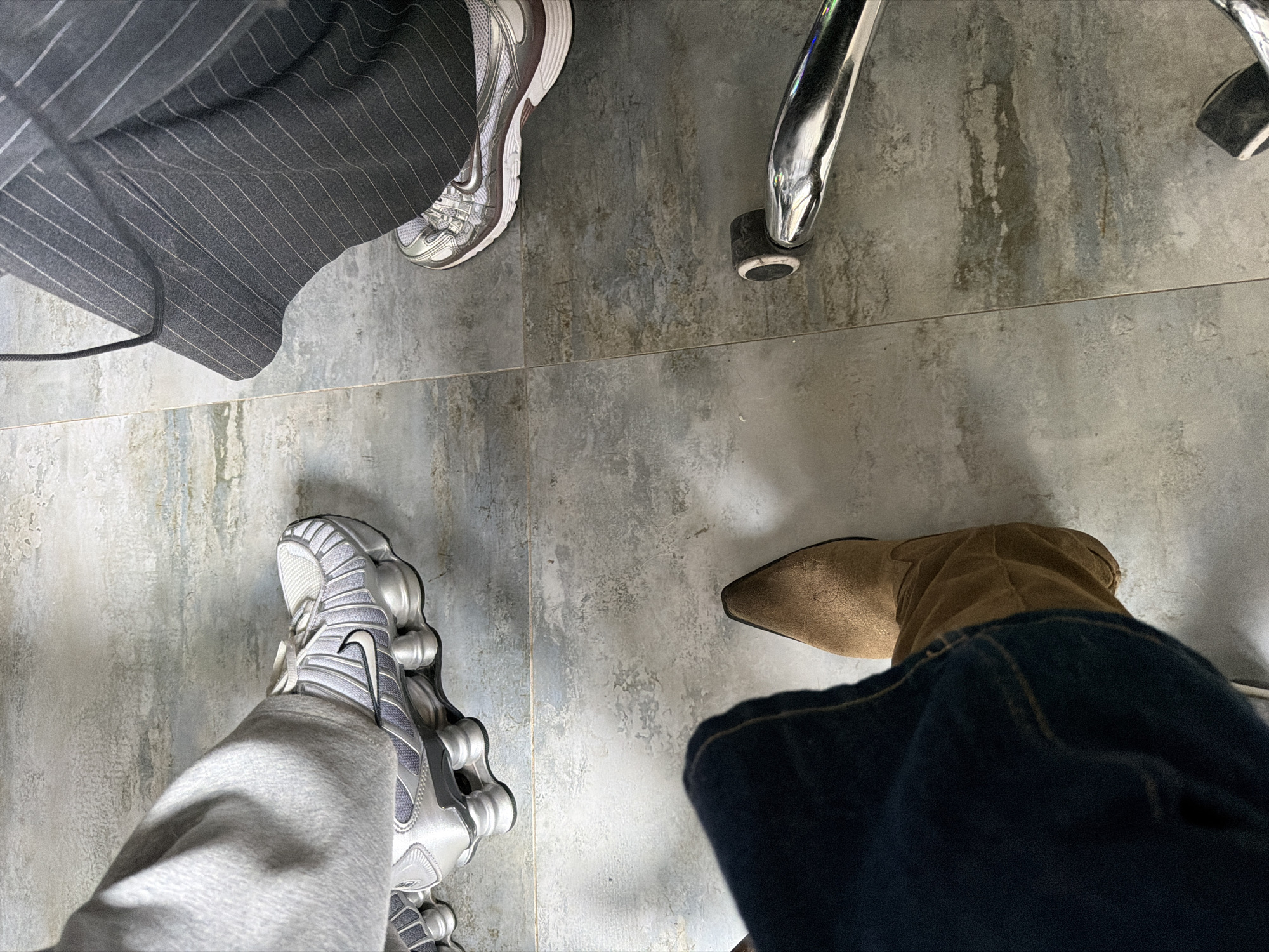 The image depicts a view of the floor from above, showing the lower legs and footwear of three individuals. The floor is tiled with large, light gray, speckled tiles. At the top left, a person is wearing gray pinstripe pants and gray and white athletic shoes. A second person is wearing gray sweatpants and silver Nike Shox athletic shoes. A third person is wearing dark blue jeans and a tan suede boot with a pointed toe. The leg of a chrome chair is also visible. The location is not discernible from the image.