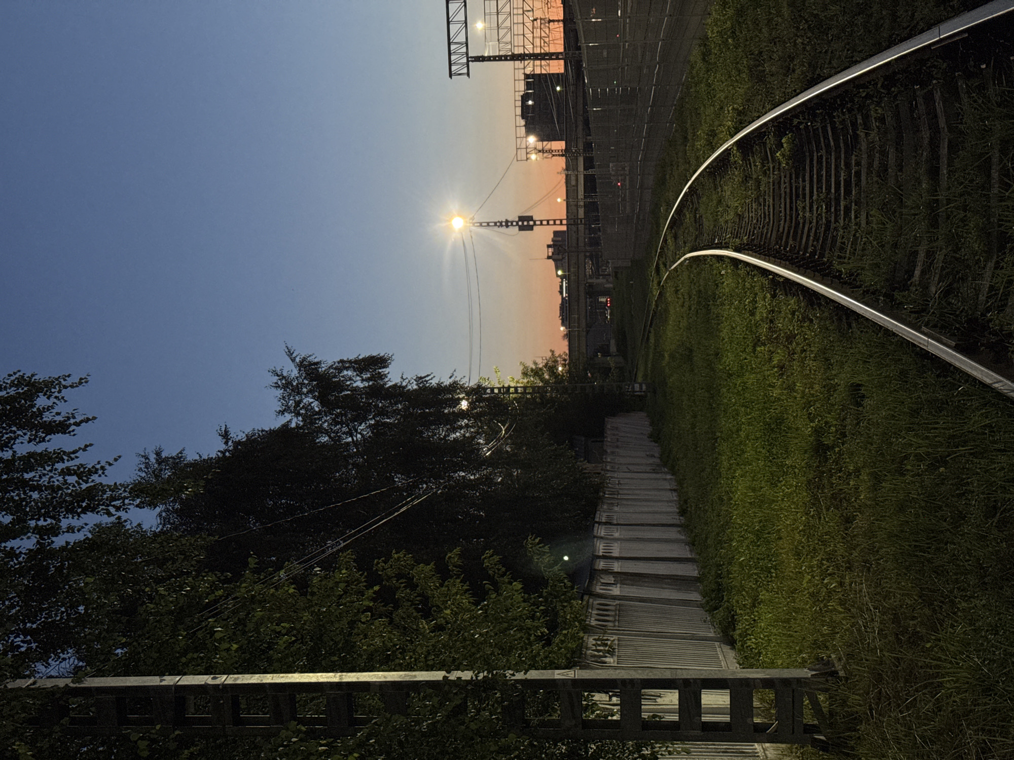 A twilight scene captures a railway track curving through a grassy embankment, flanked by a tall fence and lush trees. In the background, a street light illuminates the sky, and buildings with more lights are visible. The sky transitions from a deep blue overhead to a warm orange hue near the horizon, suggesting the aftermath of sunset. The environment is urban, possibly on the outskirts of Moscow, Russia, given the style of the railway infrastructure and buildings. No people are present in the frame.
