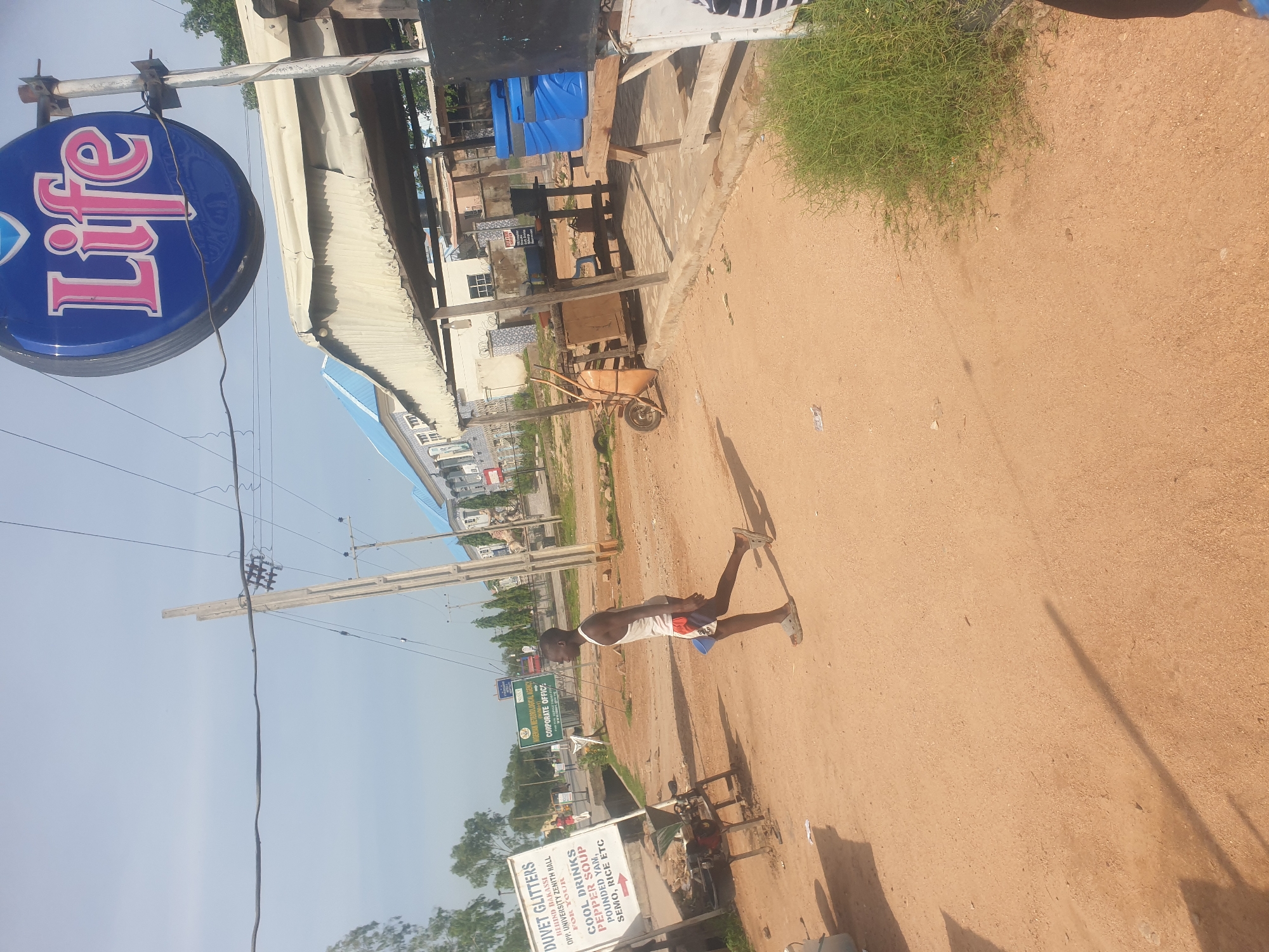 The image depicts an outdoor scene in Jauro Yinu, Nigeria. A young male, wearing a white tank top and patterned shorts, walks across a dusty, unpaved road. He is carrying a blue plastic cup in his right hand. In the background, a sign for "DUVET GLITTERS" is visible, listing items like "COOL DRINKS," "PEPPER SOUP," "POUNDED YAM," "SEMO," and "RICE." Other signs are present, including one for the "NIGERIAN METEOROLOGICAL AGENCY CORPORATE OFFICE." A blue and white "Life" branded sign hangs from a pole. The environment features utility poles with overhead wires, some scattered debris, and greenery. A covered outdoor structure with tables and chairs, possibly a vendor stall or eatery, is situated to the right. A wheelbarrow and several blue coolers are visible near the structure. The sky is clear and bright.