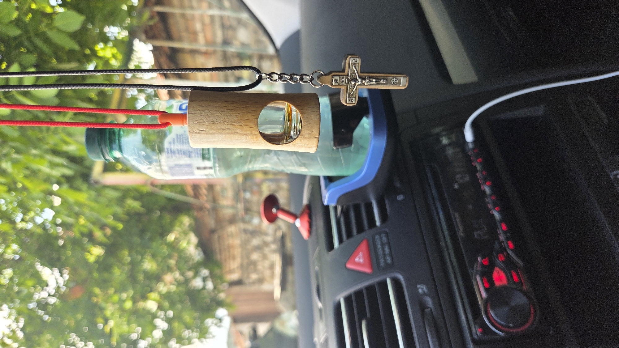 The image shows a close-up of a car's interior, focusing on various objects. In the foreground, a wooden diffuser with a glass vial is suspended by black and red cords. A small, metallic Christian cross with a chain hangs from the diffuser. Behind the diffuser, a partially visible plastic bottle of water with a green cap is held in a blue car vent clip. To the right of these items, a car's dashboard is visible, featuring a car stereo with illuminated red buttons and a dial. A white USB cable is plugged into the stereo. In the background, blurred greenery suggests an outdoor setting, possibly trees and foliage seen through the car window. The lighting indicates it is daytime, and the overall impression is of a car parked outdoors. There is no visible text in the image.