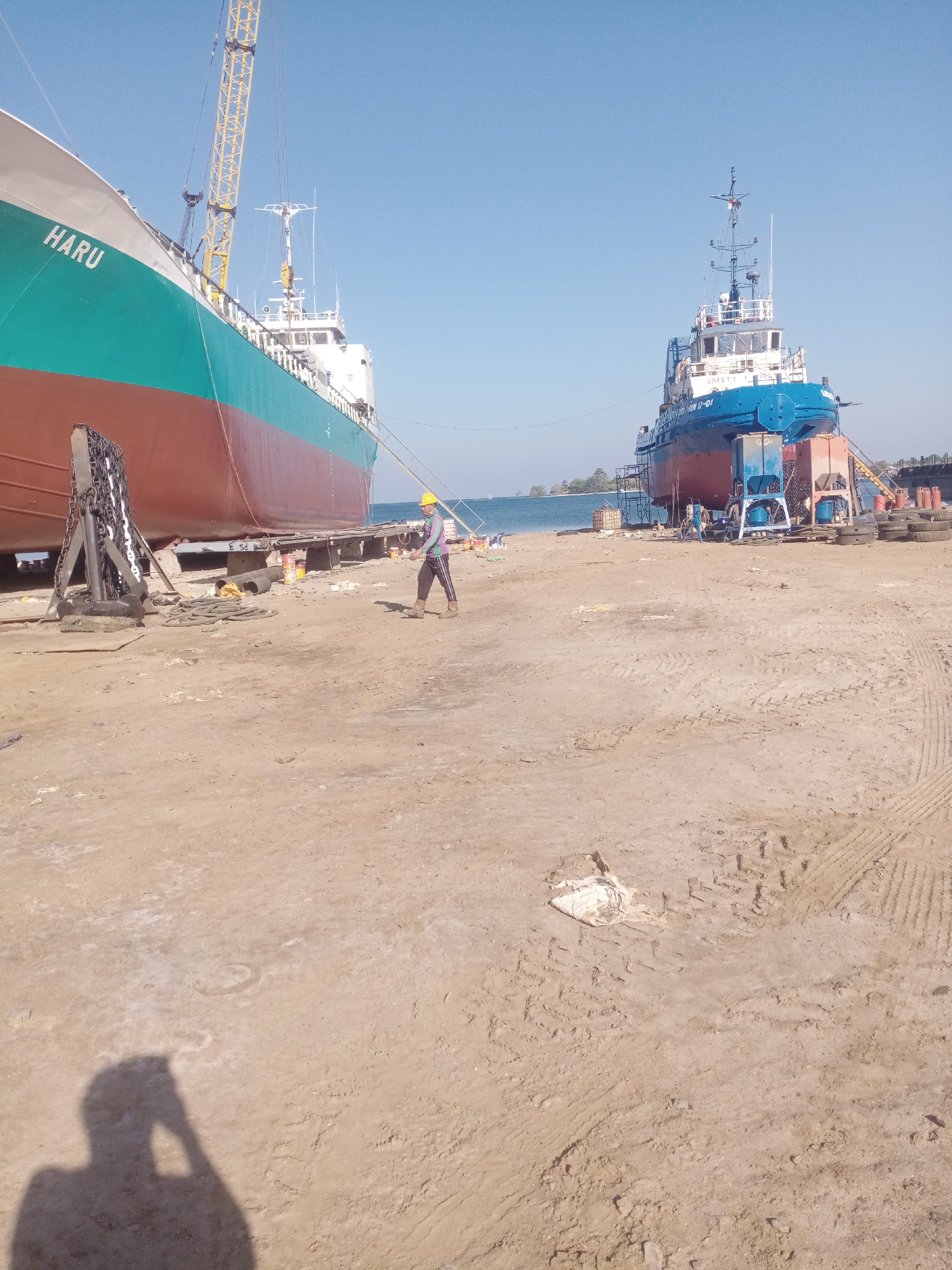 In the city of Siddo, Indonesia, the image captures a dry dock environment with two ships partially visible. The ship on the left, identified by the name "HARU," is a large cargo vessel painted with a brown hull and a turquoise upper section. To its right, a smaller blue and red tugboat stands on blocks. The ground is covered in sand and dirt, marked with tire tracks from heavy machinery. A person wearing a yellow hard hat and a light-colored long-sleeved shirt with dark pants is walking across the sandy area. In the foreground, a large anchor sits on a wooden support. The sky is clear and blue, indicating a sunny day.