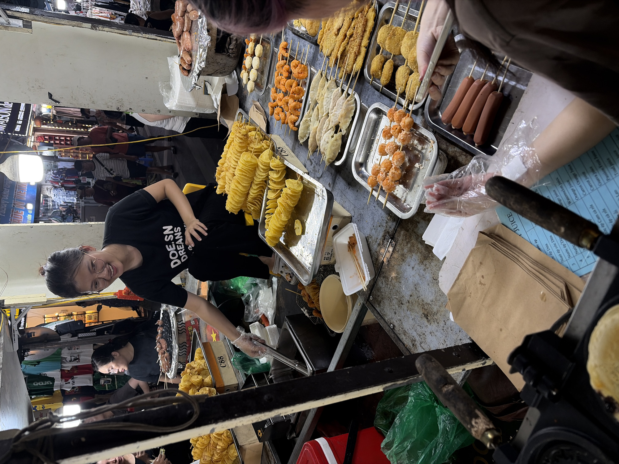 A street vendor, wearing glasses and a black t-shirt with white text reading "DOESN'T DREAM", prepares food at a market stall in Hanoi, Vietnam. She is wearing a clear plastic glove and is holding tongs. In front of her, on the stall, are several trays of food. One tray holds spiral-cut fried potatoes. Another tray has skewers of fried shrimp, and another has skewers of what appear to be dumplings or fish cakes. Further back, a larger metal pot holds a variety of sausages on skewers. Behind the vendor, other stalls are visible, with clothing and other merchandise displayed. In the background, several people are browsing the market. The overall scene depicts a bustling food stall in a lively street market environment.