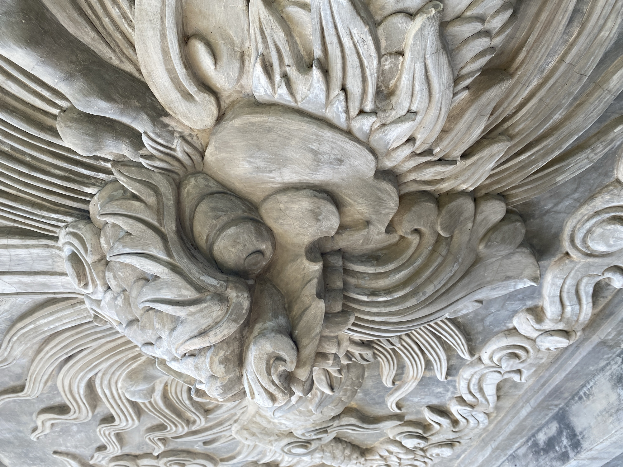 The image shows a close-up, detailed view of a stone carving of a dragon's head, likely part of a larger architectural element. The carving is intricately detailed, featuring swirling patterns, textured scales, and sharp, stylized features of the dragon's face, including its snout, teeth, and mane. The stone appears weathered, with visible cracks and variations in color, suggesting it is an outdoor structure that has been exposed to the elements. The lighting highlights the three-dimensional relief of the carving, casting shadows that accentuate its depth and texture. Based on the provided context, this carving is located in Tay Nha Trang Ward, Vietnam, suggesting it is a cultural or historical artifact within the city.
