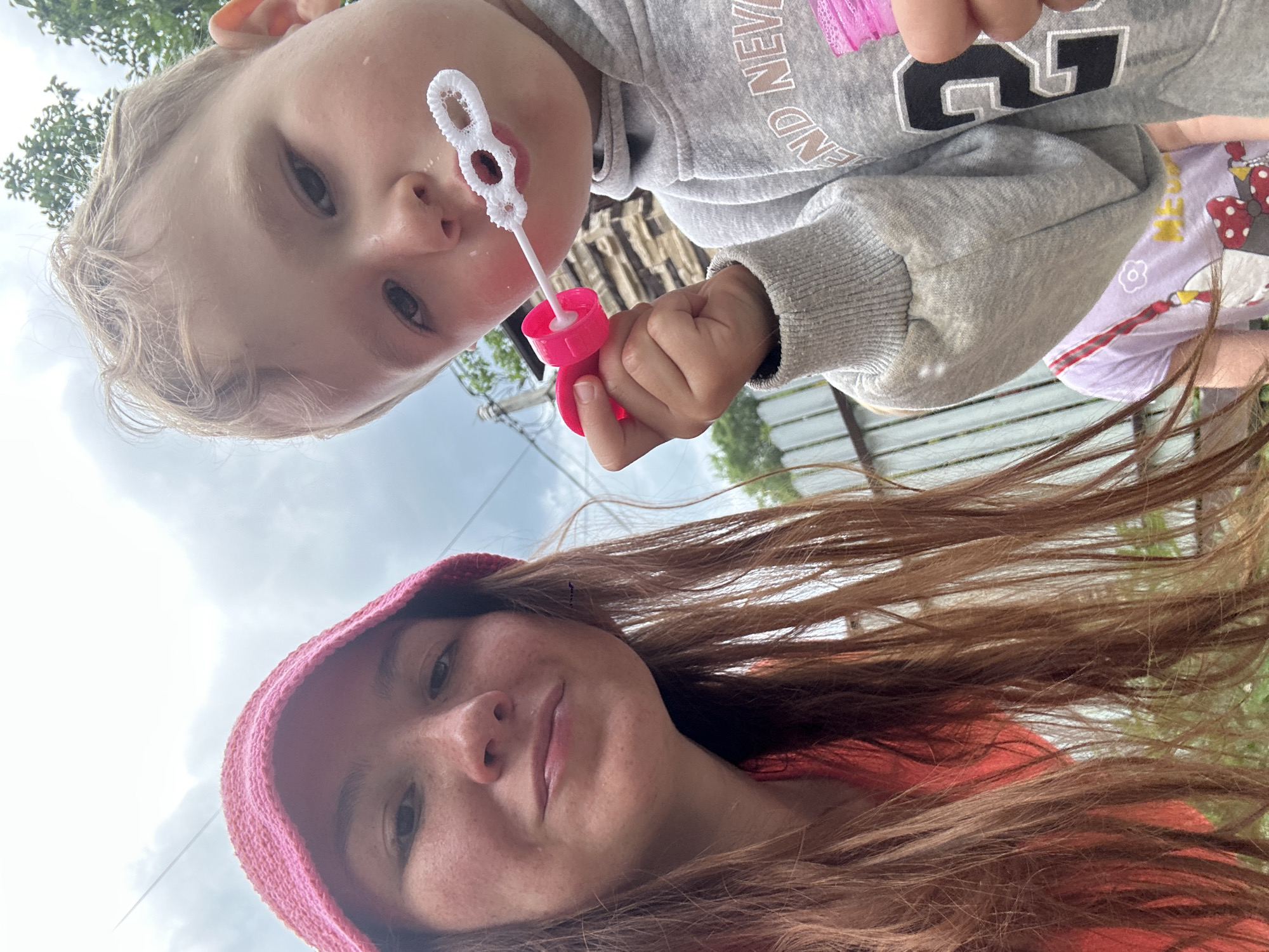 In an outdoor setting, possibly a backyard or park in Nizhny Novgorod, Russia, a young child with blonde hair is blowing bubbles with a pink bubble wand. The child is wearing a grey hooded sweatshirt with the number '2' visible on it. To the left, a young woman with long auburn hair and a pink knitted hat is looking at the camera. She is wearing an orange top. The background shows a chain-link fence and some greenery. The sky is overcast.