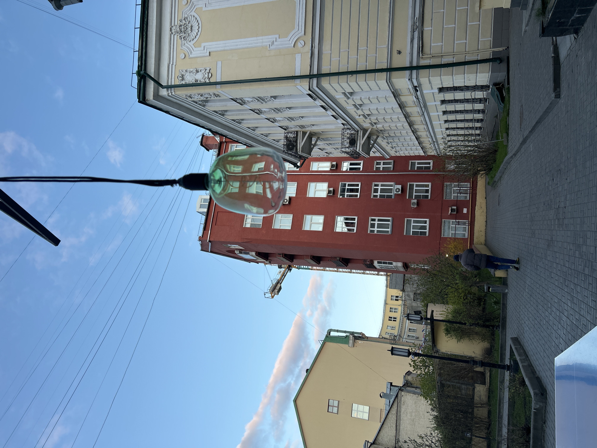 An outdoor, daytime image captures an urban scene, appearing to be an alley or courtyard between buildings. The upper portion of the frame is dominated by a clear, bright blue sky with a few wispy white clouds. Numerous thin black wires crisscross the sky.
In the foreground, slightly to the left of center, a large, clear glass light bulb hangs from a thick black wire, reflecting the red building and sky. To the right, a multi-story yellow building with ornate classical architectural details, including decorative cornices and balconies with black wrought-iron railings, is prominent. Behind this, and slightly to the left, stands a reddish-brown brick building featuring multiple windows and several visible air conditioning units. Further in the background, a lighter beige building with a green roof structure and a construction crane arm extending into the sky can be seen.
On the lower right, a paved pedestrian path made of grey interlocking pavers curves into the distance. A single person, dressed in a dark jacket and blue pants, walks away from the viewer on this path. Scattered green vegetation, possibly small trees or bushes with sparse leaves, lines the path, suggesting late autumn or early spring. A black streetlamp post is also visible along the path. The scene conveys a calm, everyday urban environment. No discernible text is visible in the image.