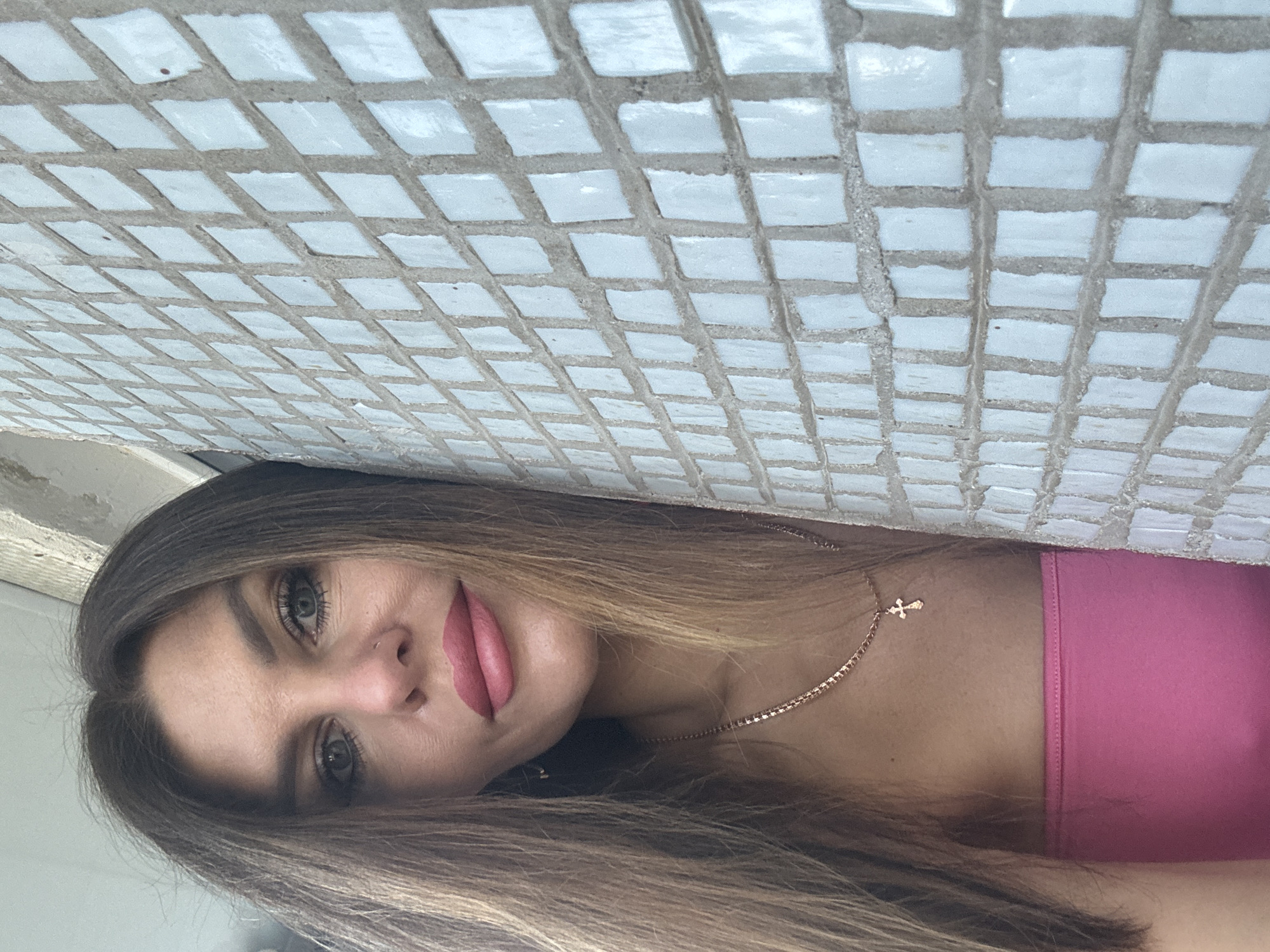 A woman with long, brown hair and blue eyes is leaning against a wall tiled with small, square, light blue tiles. She is wearing a pink strapless top and a gold necklace with a cross pendant. Her makeup includes pink lipstick and eyeliner. The wall is the primary background element to her right, and the background to her left is out of focus. The image appears to be a selfie, taken from a slightly upward angle. The specific location in Chaikovsky, Russia, cannot be determined from the visual information provided.
