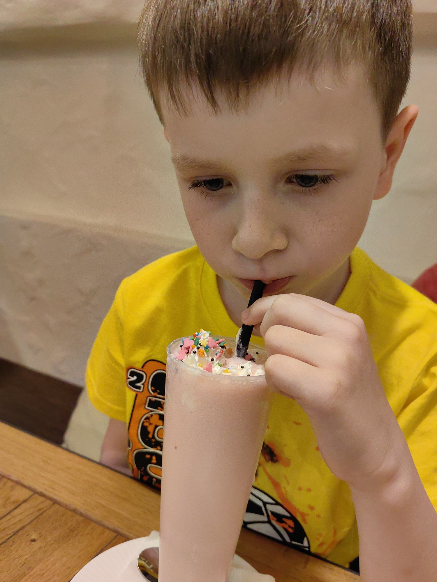 A young, light-skinned child with short, light brown hair and visible freckles on the nose and cheeks is depicted in an indoor setting, located in Sochi, Russia. The child, estimated to be pre-adolescent, is wearing a bright yellow t-shirt with a partial orange and black graphic on the viewer's left side. The child's gaze is directed downwards, focused on a tall, clear glass held by the right hand. The glass contains a pale pink, opaque beverage, consistent with a milkshake or smoothie. The drink is topped with white whipped cream, an assortment of small, multicolored confectionery sprinkles (including red, blue, green, yellow, white, and orange), and small white decorative candies. A black straw is inserted into the drink, positioned between the child's lips, indicating consumption of the beverage. The child's right hand is grasping the upper portion of the glass. The foreground features a wooden table with visible grain and paneling. The background consists of a uniformly light-colored, possibly beige or cream-toned, textured wall, out of focus, characteristic of an interior space such as a restaurant or cafe. No specific architectural or geographical identifiers are discernible within the image to confirm the location visually.