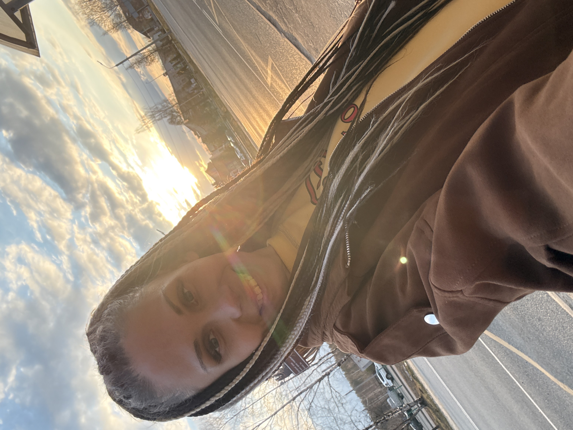 A person is photographed outdoors at eye level, angled upwards, with the sunset visible in the background. The person, likely female, is smiling with braided hair. She is wearing a brown jacket over a yellow top. The background features a street with lane markings and buildings under a cloudy sky with the sun setting on the horizon. The photograph appears to be taken in a residential or urban area. There is no direct confirmation that the location is in the city of Rusanovo, Russia based on visual information alone.