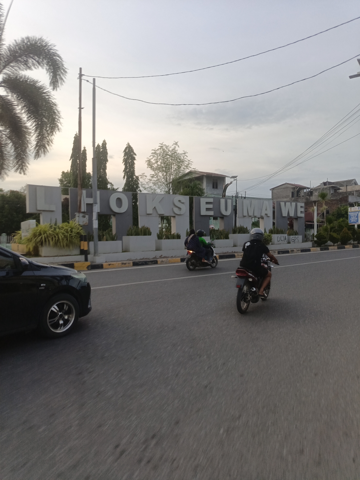 The image shows a view of a street in Lhokseumawe, Indonesia, with a large sign in the background that reads "LHOKSEUMAWE". The sign is made of large, grey, block letters and is situated behind a line of manicured bushes and plants. Two individuals are seen riding motorcycles on the asphalt road. The motorcycle in the foreground is being ridden by a person wearing a black shirt and a helmet, with another person on the back also wearing a helmet. The motorcycle in the background has two people on it, one driving and one passenger. A black car is partially visible on the left side of the image, moving along the road. The sky is overcast, suggesting it is either early morning or late afternoon. Power lines are visible stretching across the sky. Several buildings, some appearing to be houses or small businesses, are visible behind the "LHOKSEUMAWE" sign. The road has yellow and black stripes along the edge.