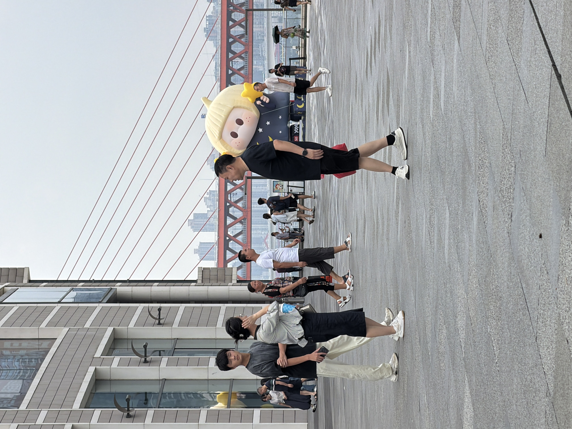 The image depicts a street scene in Yuzhong District, China, on a cloudy day. In the foreground, a man in a black t-shirt and shorts walks towards the viewer's left. Behind him, a large inflatable character resembling a child with blonde hair and holding a yellow star is visible. To the left, a young man in a gray t-shirt and light-colored pants stands with his phone in his hand, facing forward. Beside him, a woman in a striped shirt and dark skirt touches her hair. Other pedestrians are scattered throughout the scene, walking in various directions. In the background, a modern building with large windows is visible, along with a bridge with red suspension cables spanning across a body of water. The ground is wet, suggesting recent rain.