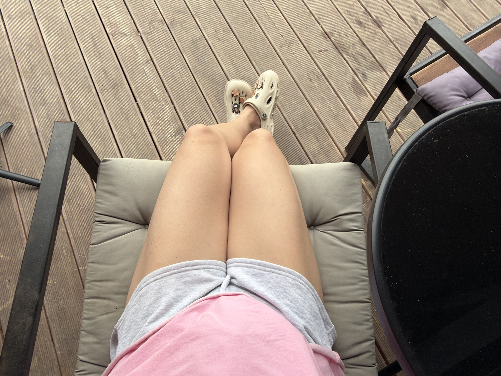 The image shows a person's legs from the knees down, wearing a pink t-shirt and grey shorts. They are sitting on a cushioned chair, which is placed on a wooden deck. The person is also wearing cream-colored Crocs with charms. To the left, a portion of a black table is visible. The scene appears to be outdoors, possibly on a patio or balcony, with wooden planks making up the floor. The location is identified as Семилуки, Russia.
