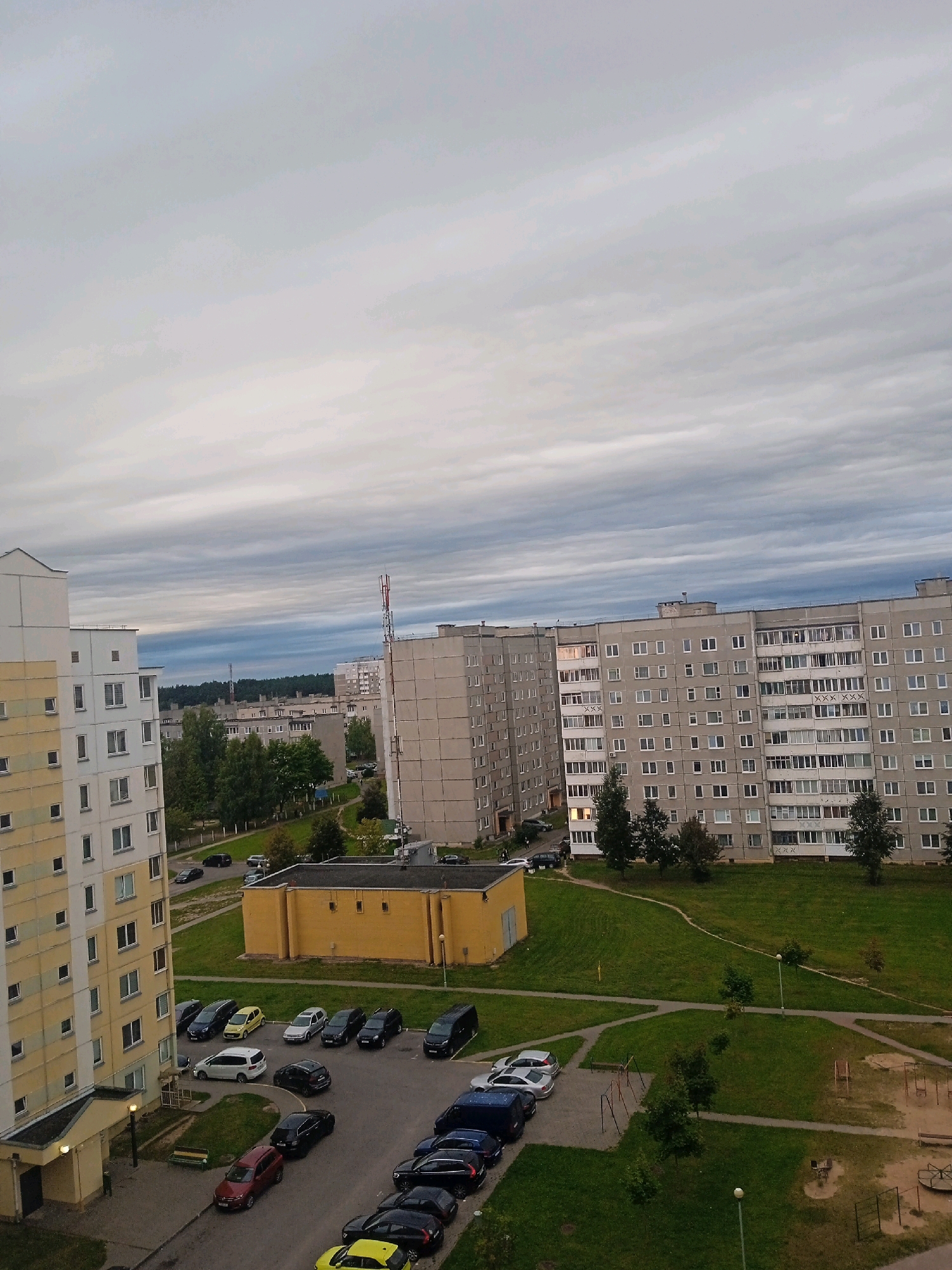 The image captures a wide-angle, elevated view of a residential area in Lida, Belarus. In the foreground, a tall, light-yellow apartment building with numerous windows dominates the left side. To the right, several larger, multi-story gray apartment buildings are visible, characterized by their numerous windows and balconies. A prominent yellow rectangular building with a dark roof is situated in the middle ground, bordered by a manicured lawn.
The scene includes several cars parked in designated areas and along roads. A variety of vehicles, including sedans, vans, and SUVs, are parked in the driveways and parking lots of the apartment buildings and near the yellow structure. Green grassy areas and walking paths connect the buildings. In the distance, a forest line can be seen under a cloudy sky. The overall impression is of a typical Soviet-era residential complex. There are no visible people in the image.