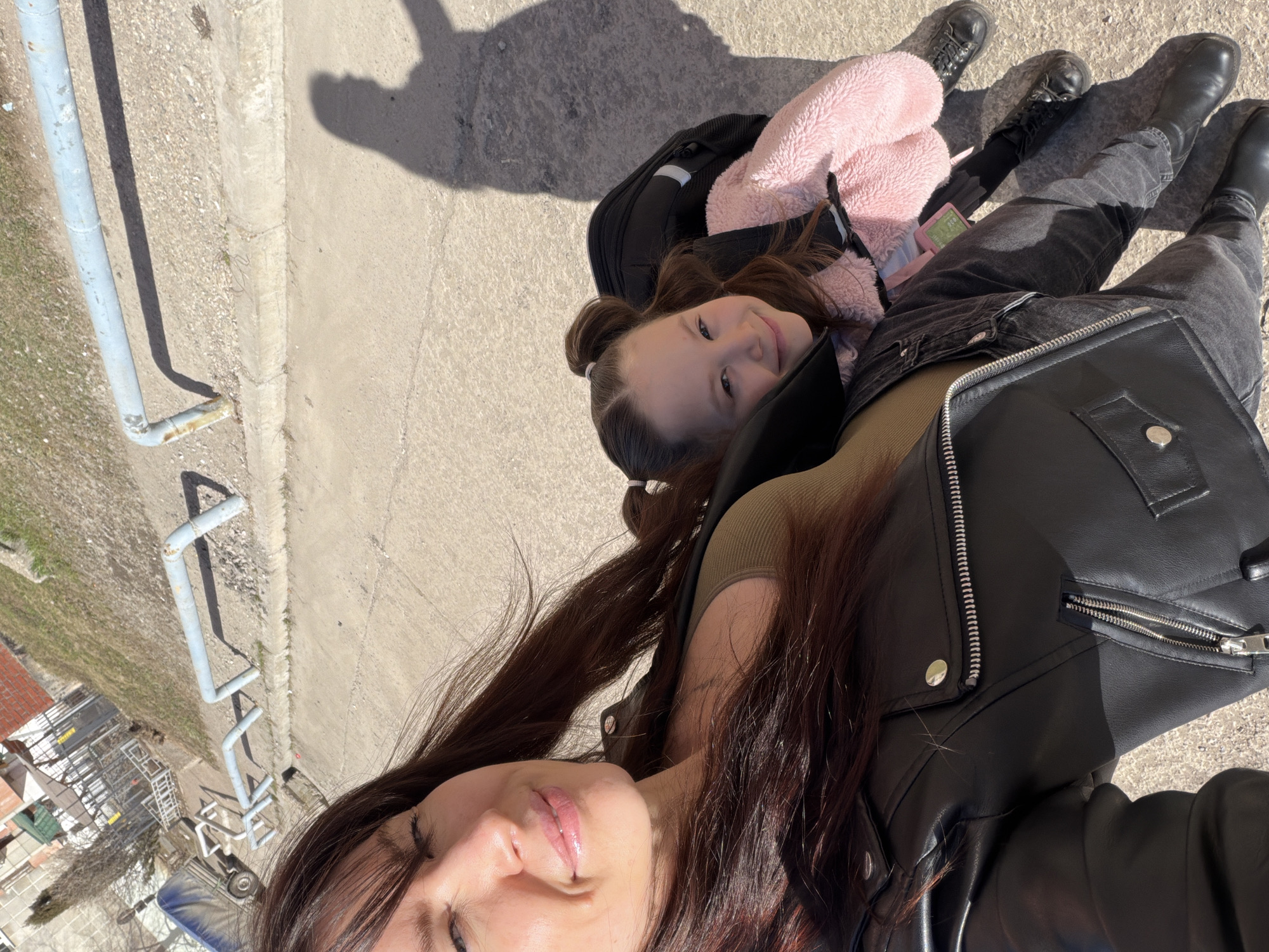 A selfie photograph, taken in daylight, shows a woman and a young girl in an urban environment. The woman, in the foreground, is partially visible from the chest up, wearing a black leather jacket and a muted green top. She has dark brown hair. The girl is lying behind her and to the left, also partially visible. She is wearing a pink fuzzy jacket and black pants, and has her hair pulled back in two small buns. Both individuals are facing the camera with slight smiles. The background shows a concrete surface and a section of scaffolding, and possibly a street or sidewalk. The photo was taken outside, as indicated by the bright sunlight and shadows. Without further information, I cannot confirm it was taken in Perm, Russia.