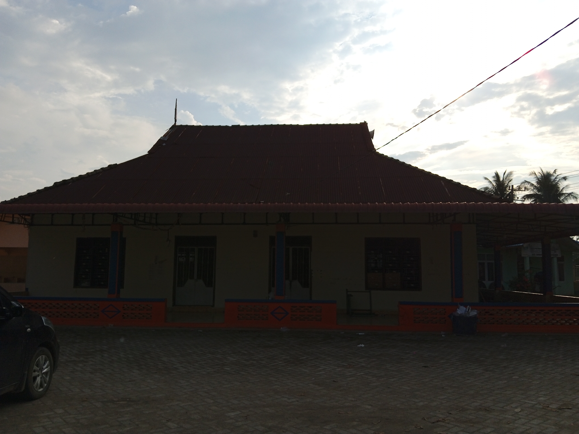 The image shows a low-angle exterior shot of a building with a red corrugated metal roof, located in Cotpuuk, Indonesia. The building has a covered porch area with orange decorative fencing along the bottom. Several windows and doors are visible on the building's facade. To the left, the front portion of a dark-colored car is parked on a paved surface. The sky above is partly cloudy with the sun visible, suggesting it is daytime. Palm trees are visible in the background to the right. The overall scene appears to be a residential or community building in a tropical or subtropical environment.