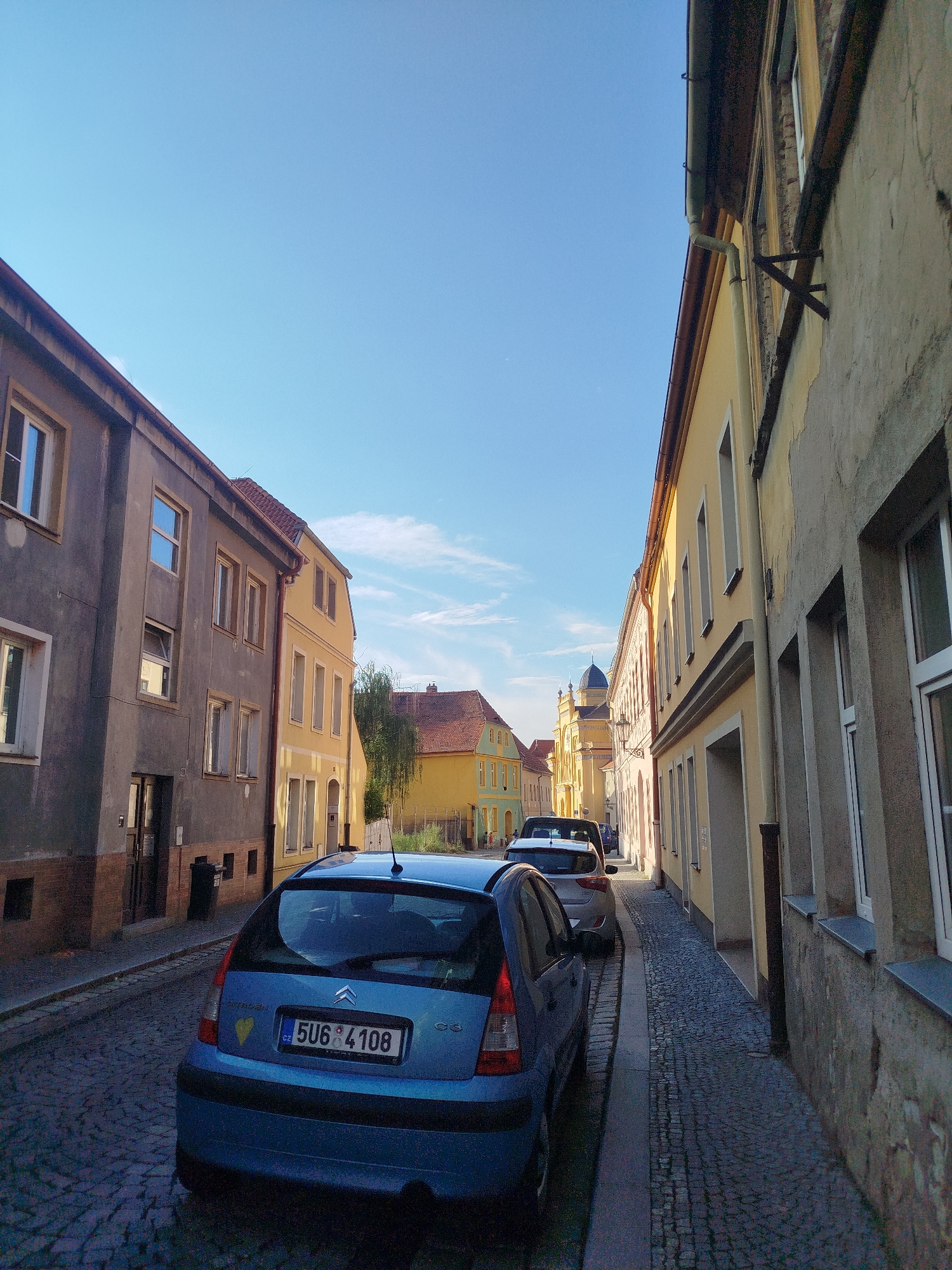 This media file depicts a narrow street lined with old European-style buildings under a clear blue sky with scattered clouds. The architecture suggests a historic European town. On the left, a gray building with several windows stands tall. To its right, a yellow building with more windows follows the street's curve. On the right side of the street, another yellow building with a textured, aged facade is visible, with several windows and architectural details.
The street itself is paved with cobblestones. Several cars are parked along the street. In the foreground, a blue hatchback car is parked, facing away from the camera. Its license plate reads "CZ 5U6 4108". Further down the street, more cars are visible, including a white SUV.
The scene appears to be set during daytime, likely late afternoon or early evening, given the angle of the sunlight and the warm tones on the buildings. The sky is bright and clear, indicating fair weather. The overall mood is peaceful and picturesque, capturing a quiet moment in a charming old town. The presence of buildings with classic European architectural features, including tiled roofs and a distinct dome-shaped structure visible in the distance, further reinforces the European location. There is no overt activity or event taking place; the scene is a static view of the street.
