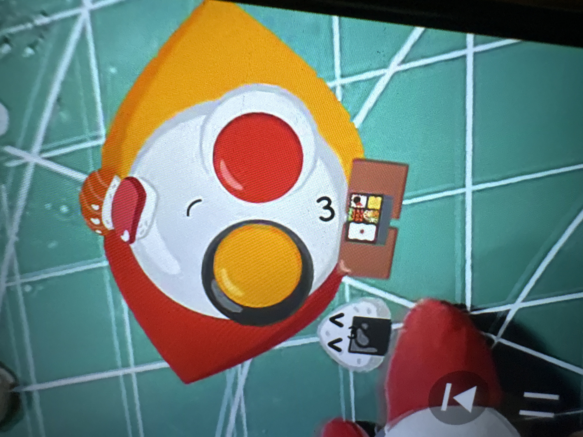 The image shows a close-up, low-angle view of a video game or application interface displayed on a screen. The main subject is a cartoonish, anthropomorphic character resembling a sushi roll with a happy face. The character has a round white head with a curved black line for a mouth, and two large circles for eyes: one orange and one red. A red and white piece of sushi sits on top of its head. The character is wearing a red and orange outer garment, possibly representing a stylized kimono or cape. Below the character's head is a small brown platform displaying what appears to be a bento box with various food items. To the left of the main character is a small, triangular onigiri (rice ball) character with a black seaweed wrapper and a smiling face with large eyes. To the far left of the frame, a red and white object with a play button icon is partially visible. The background is a teal-colored grid pattern, possibly representing a game board or interface element. The lighting suggests the image is taken in a dimly lit environment, likely indoors, reflecting off the screen. There is no visible indication of Ashvem, India, or any real-world city location.