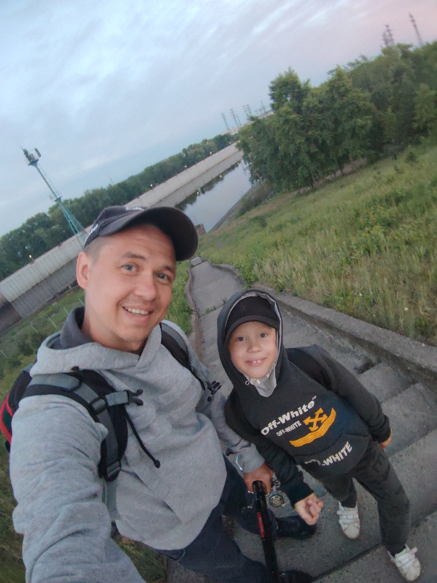 This selfie captures an adult man and a young boy, likely father and son, smiling brightly outdoors in Chaikovsky, Russia. The man, on the left, wears a grey hoodie, a black baseball cap, and a backpack. The boy, on the right, wears a dark grey/black hoodie with the hood up, a black baseball cap underneath, dark pants, and white sneakers. His hoodie prominently displays the text "Off-White™ OFF-WHITE" with a yellow boat-like logo featuring a black 'X', and "WHITE" printed below. Both individuals are holding a fishing rod, suggesting they are on a fishing excursion.
They are positioned on a concrete embankment or steps leading down to a calm body of water, likely a river or canal. A grassy slope rises on the right, and a line of trees borders the far bank of the water. The sky is overcast, casting a soft, even light over the scene. In the background, beyond the trees, several industrial or utility structures, including a tall metal mast and power line towers, are visible. The overall mood conveyed by their smiles is happy and content, capturing a shared moment during an outdoor activity.