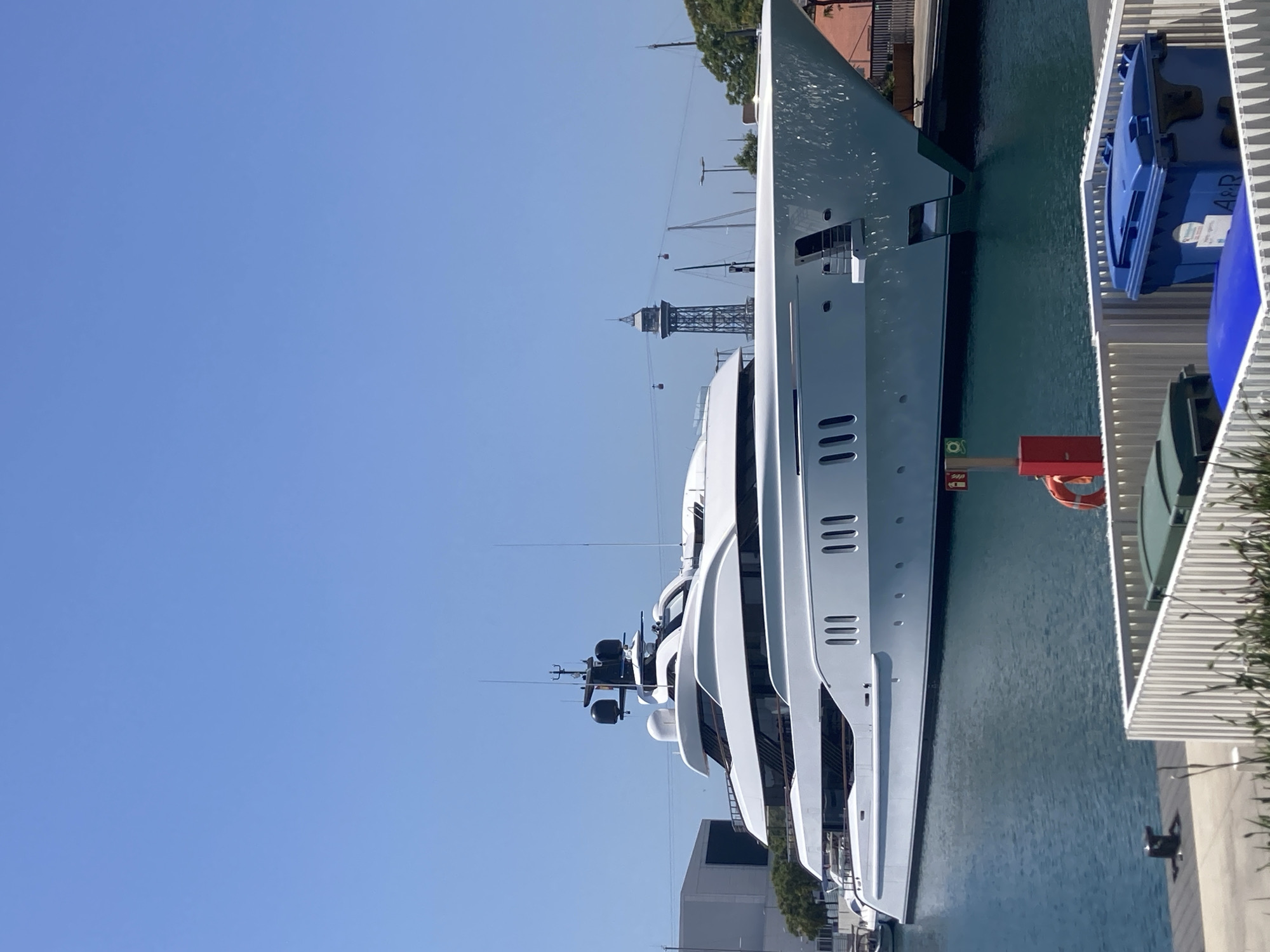 A large, modern white superyacht is docked in a harbor under a clear blue sky. The yacht has multiple decks with sleek, angular lines and numerous windows. In the background, there are port structures, including cranes and a distinctive lattice tower with a cable car system. Trees and buildings are also visible along the waterfront. To the right of the yacht, a white railing separates the dock from a waterfront promenade. A blue garbage bin with the text "A&R" is visible on the promenade, along with a red life preserver hanging on a post. The scene suggests a sunny day, possibly in the late morning or early afternoon, given the bright light and clear sky. The overall impression is one of luxury and a busy, modern port environment. The location context indicates this is likely Barcelona, Spain.