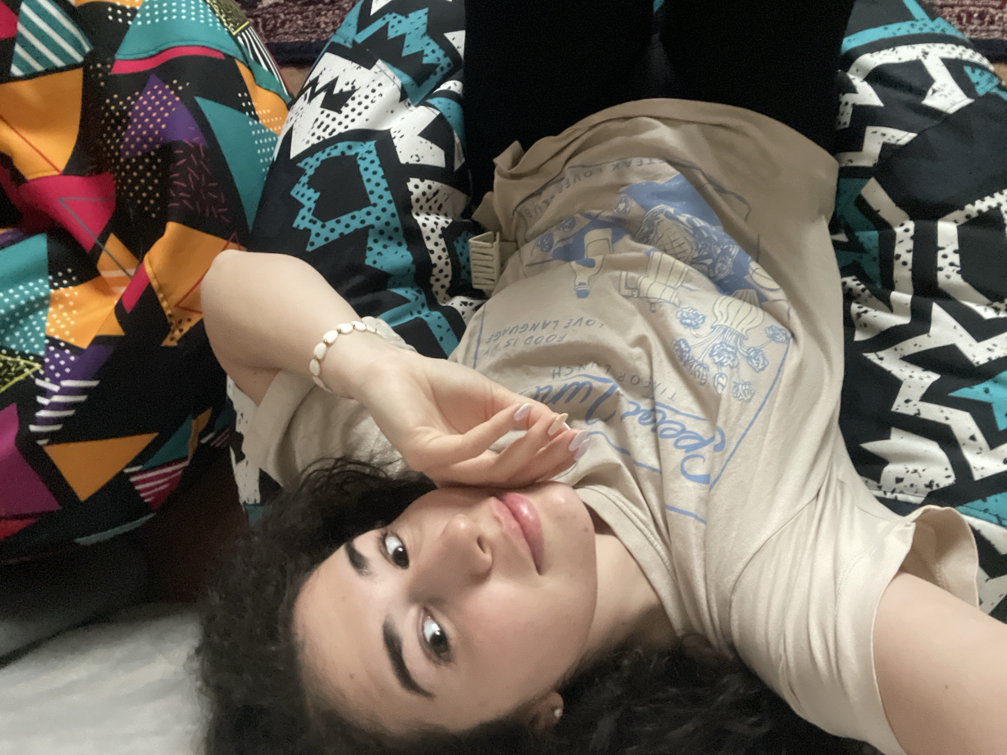 A young woman with dark curly hair is lying down, possibly on a bed or soft surface. She is facing the camera with her head tilted slightly to the right, and her hand is raised near her face. She is wearing a beige t-shirt with blue lettering and graphics. A white beaded bracelet adorns her wrist. The background includes colorful patterned fabrics or cushions. The location is not discernible from the visual information provided in the image. There is no way to tell this image was taken in Sofia, Bulgaria.