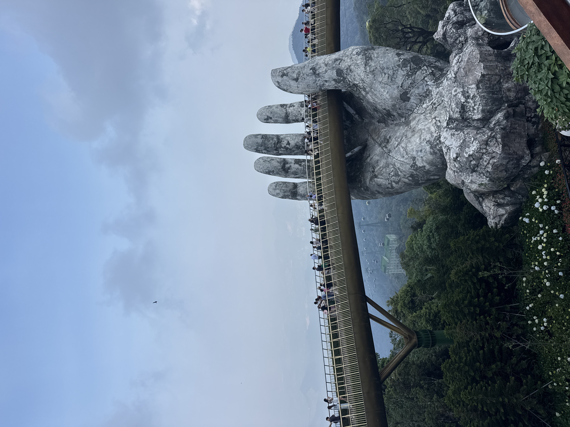 The image shows the Golden Bridge in Đà Nẵng, Vietnam, supported by two large, weathered stone hands. The bridge is crowded with people walking along its length. Lush green trees and vegetation surround the bridge. In the background, there are mountains and glimpses of buildings. The sky is overcast with gray and blue tones. A small bird flies through the sky.