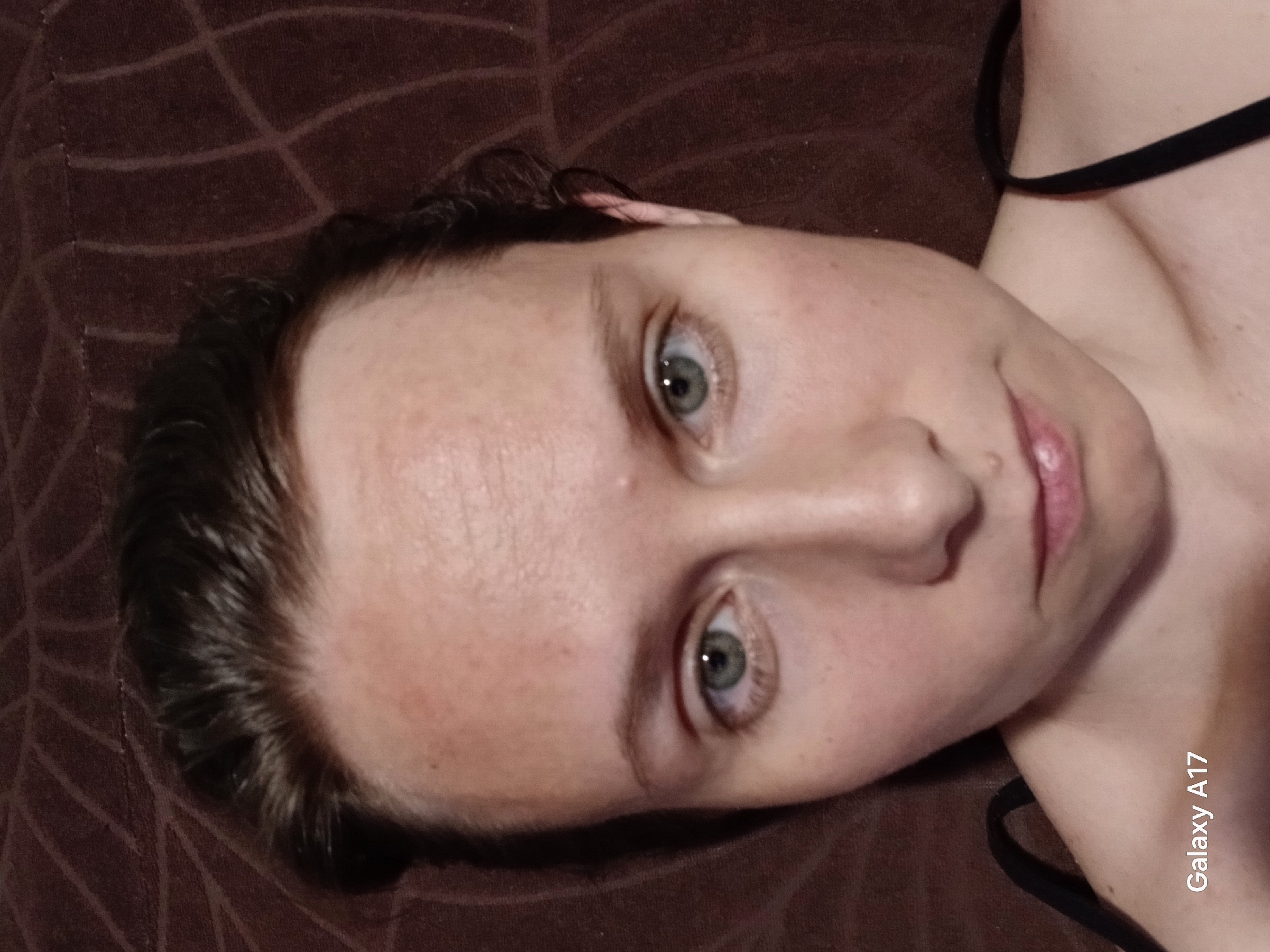 A close-up, high-angle shot shows a light-skinned person with brown hair and green eyes. The person is lying down on a brown textured surface. The person is wearing a black strap top. The image includes a "Galaxy A17" watermark on the bottom right, indicating the image may have been taken with a Samsung Galaxy A17. There is no indication of the location being Yekaterinburg, Russia.