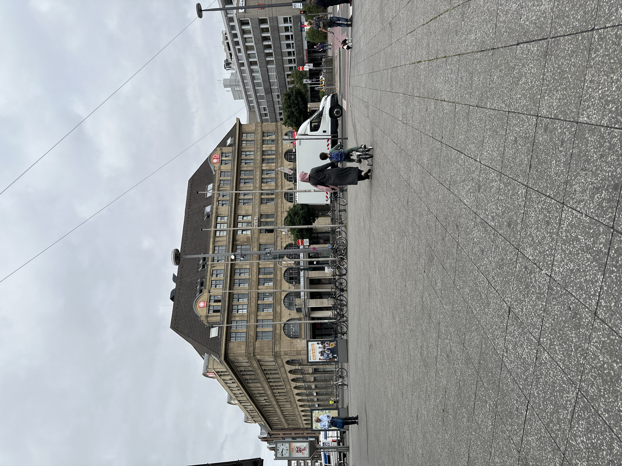 The image depicts a city square in Krefeld, Germany. In the background, a large, historic building with a mansard roof and arched windows dominates the scene. Above the arches, red logos are visible on the facade. To the right of this building is a more modern, multi-story building with a grey facade and numerous windows.
In the foreground, a paved square is occupied by several people. A woman wearing a pink hijab and a long, dark coat walks with a young child on a small bicycle. The child wears a backpack. Further to the right, a person walks a dog on a leash. The pavement is made of large, grey square tiles. Various bicycles are parked along the side of the building in the background. A white delivery van is also visible in the middle ground. The sky is overcast and grey.