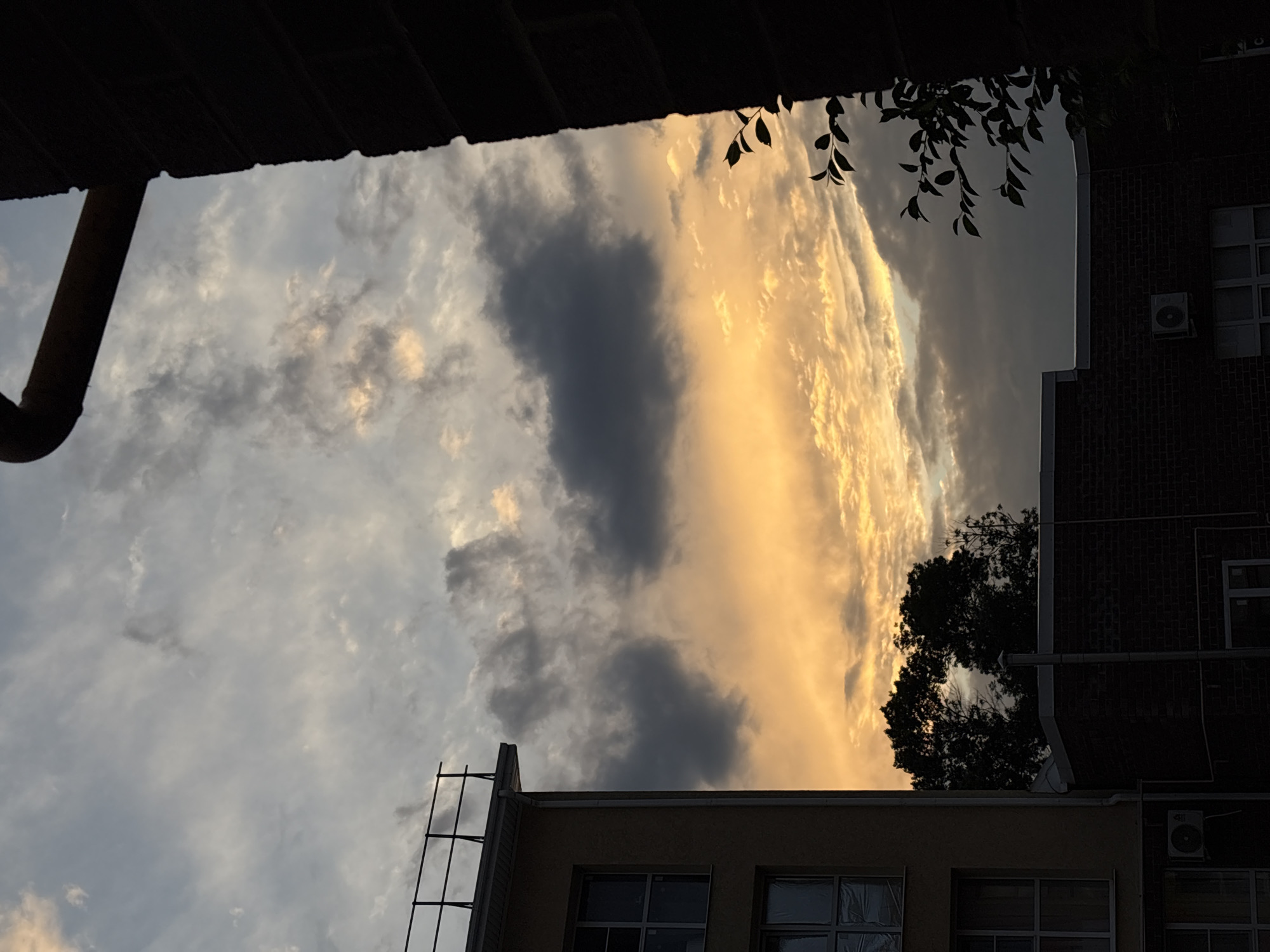 The image captures a dramatic sky scene, likely at sunset given the golden hues illuminating the clouds. The sky is filled with a mix of dark, heavy clouds and streaks of bright, warm light, creating a striking contrast. Below the sky, architectural elements are visible, including the dark, shadowed roof of a building in the foreground with a pipe extending from it. To the right, a dark brick building rises, its upper floors showing windows and an outdoor air conditioning unit. A lighter-colored building with windows is partially visible at the bottom, featuring a metal structure resembling a ladder or scaffolding attached to its side. Silhouetted trees are also present, adding to the layered composition. The scene suggests an urban or suburban setting, with buildings framing the expansive sky. There is no visible text in the image.
