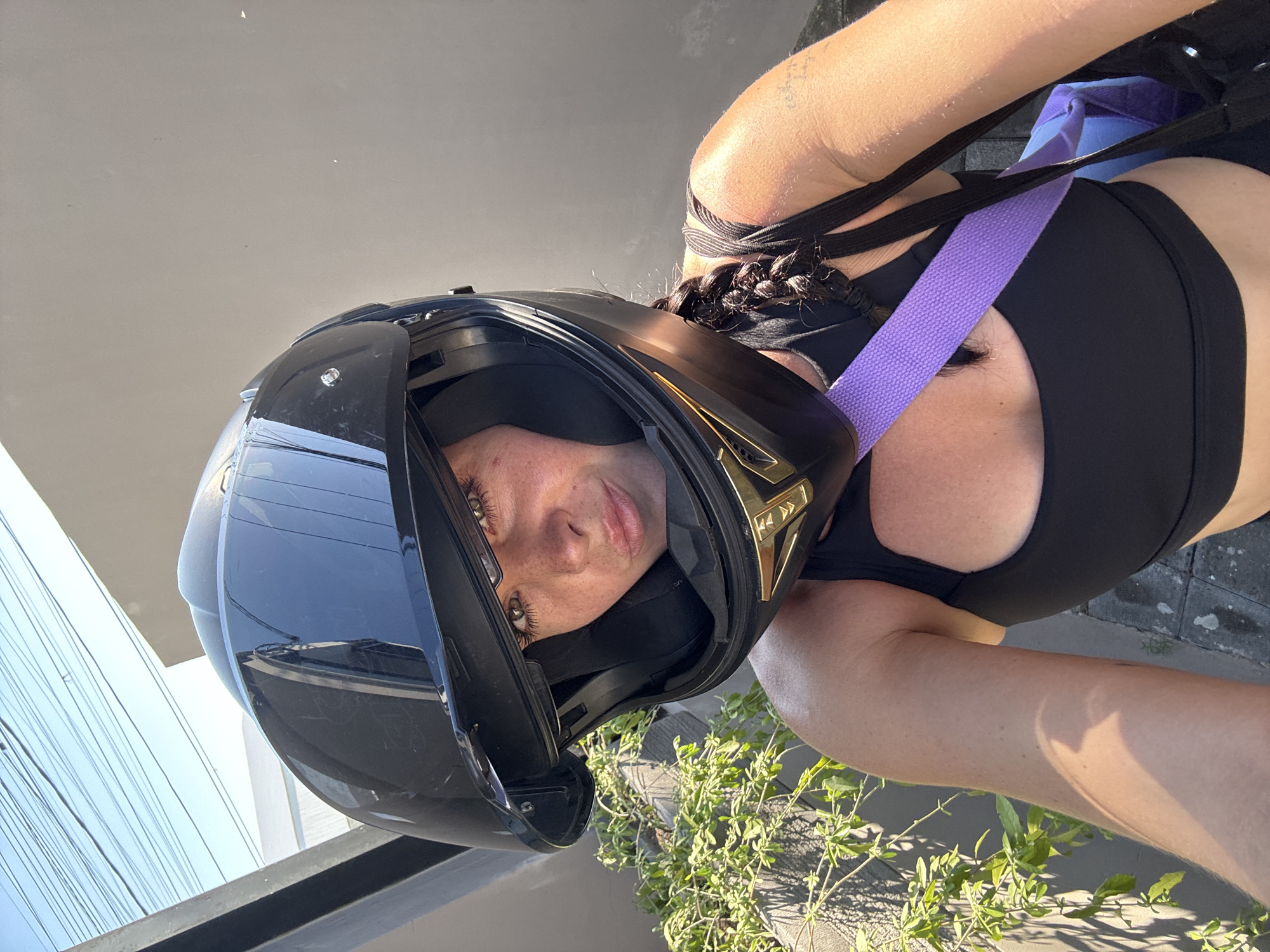 A woman is taking a selfie outdoors, wearing a black motorcycle helmet and a black athletic top. She has her hair in a braid. A purple strap from a bag is visible across her chest. There is a potted plant to her left and a grey wall behind her. The sky is visible above with power lines. The location is North Kuta, Indonesia.
