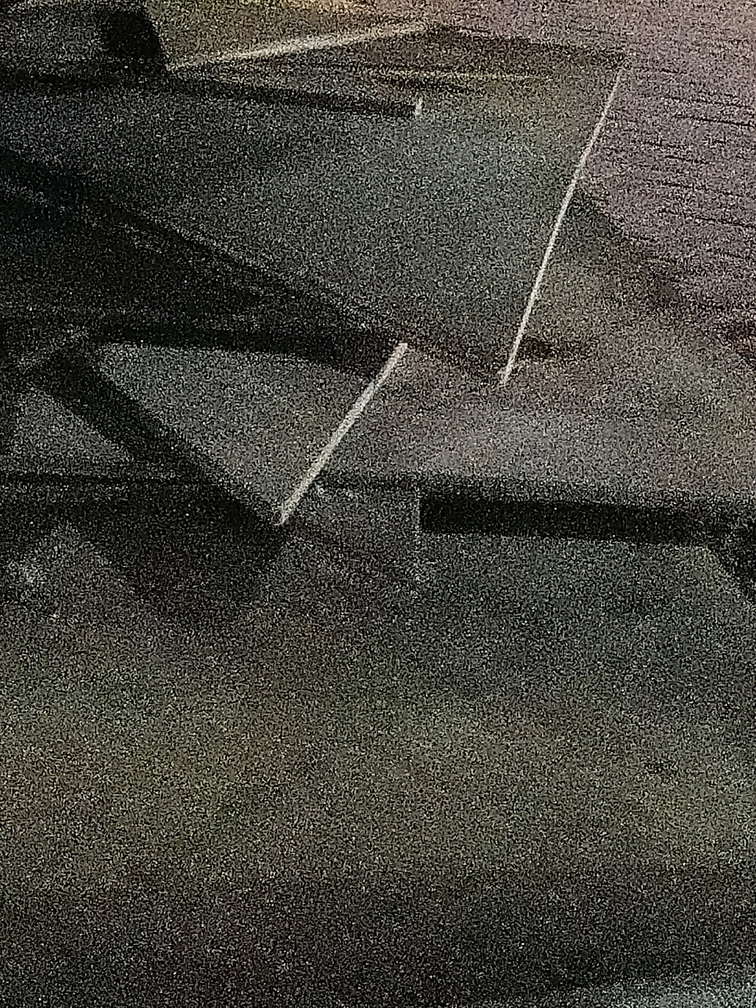 The image is a low-resolution, grainy photograph taken from an elevated perspective looking down at a dark, possibly metallic surface. The surface appears to be a rooftop or a walkway with several distinct geometric shapes and lines formed by the arrangement of these elements. There are angled planes and sharp edges visible, suggesting a modern architectural design or possibly debris. A thin, reflective white line cuts diagonally across the upper right portion of the image. The overall color palette is muted, with shades of dark grey and black dominating, interspersed with some lighter, potentially reflective areas. The image does not contain any recognizable landmarks or features that would allow for identification of its location within Cikal, Indonesia.